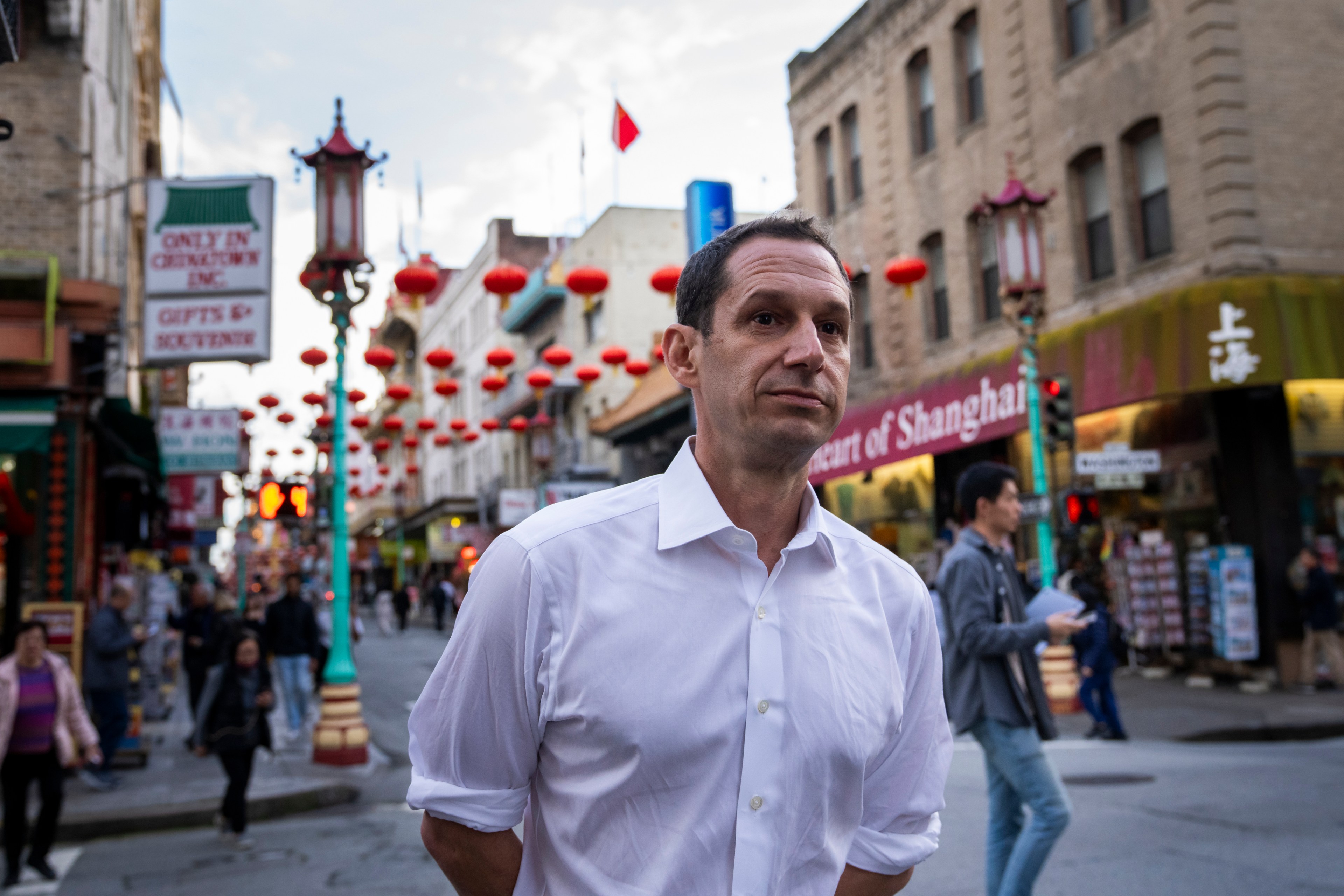 A man in a white shirt stands on a bustling street decorated with red lanterns, likely in a Chinatown. People walk by, and signs advertise gifts and Shanghai.