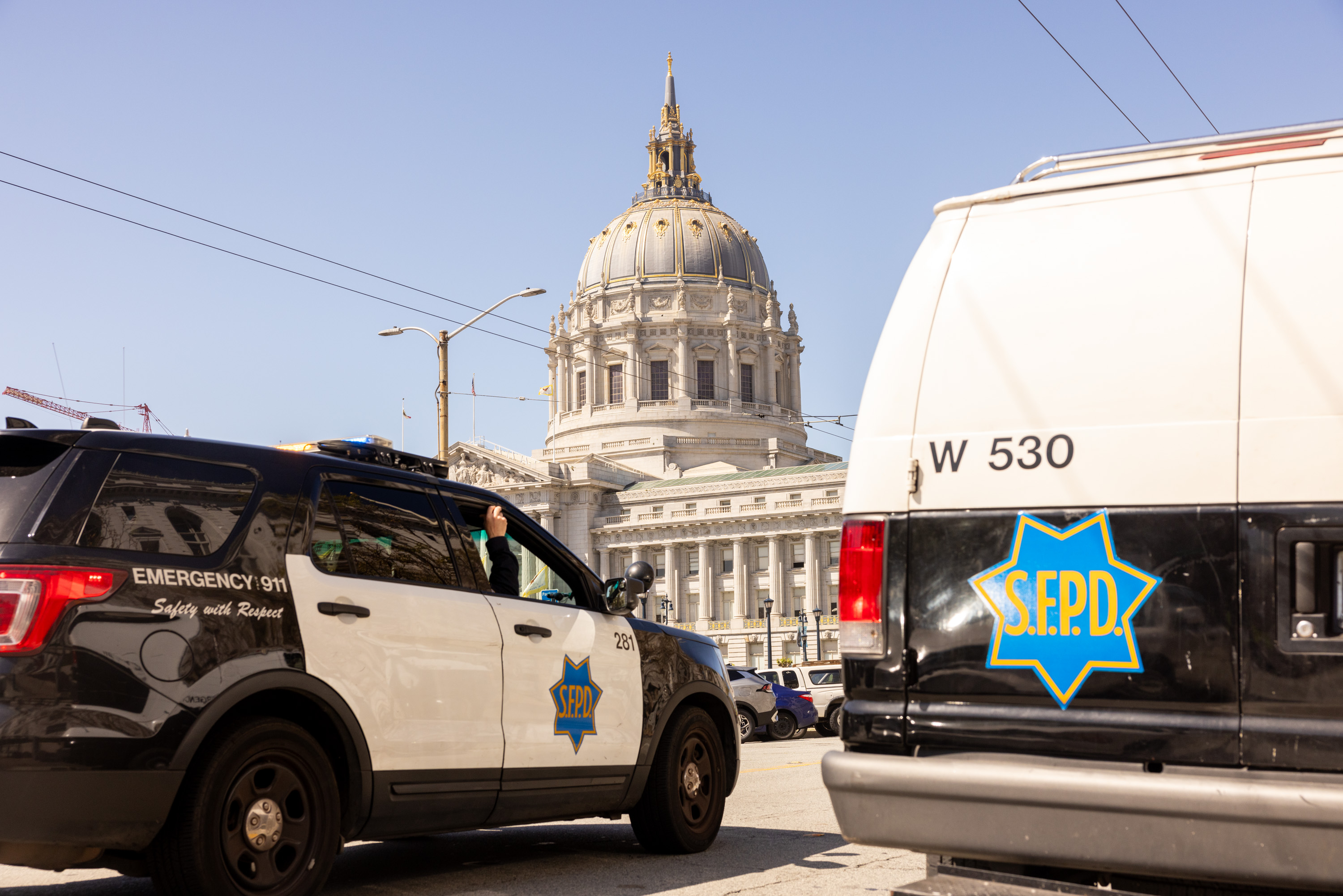 A police SUV and van with S.F.P.D. logos are parked near a grand domed building under a clear blue sky.