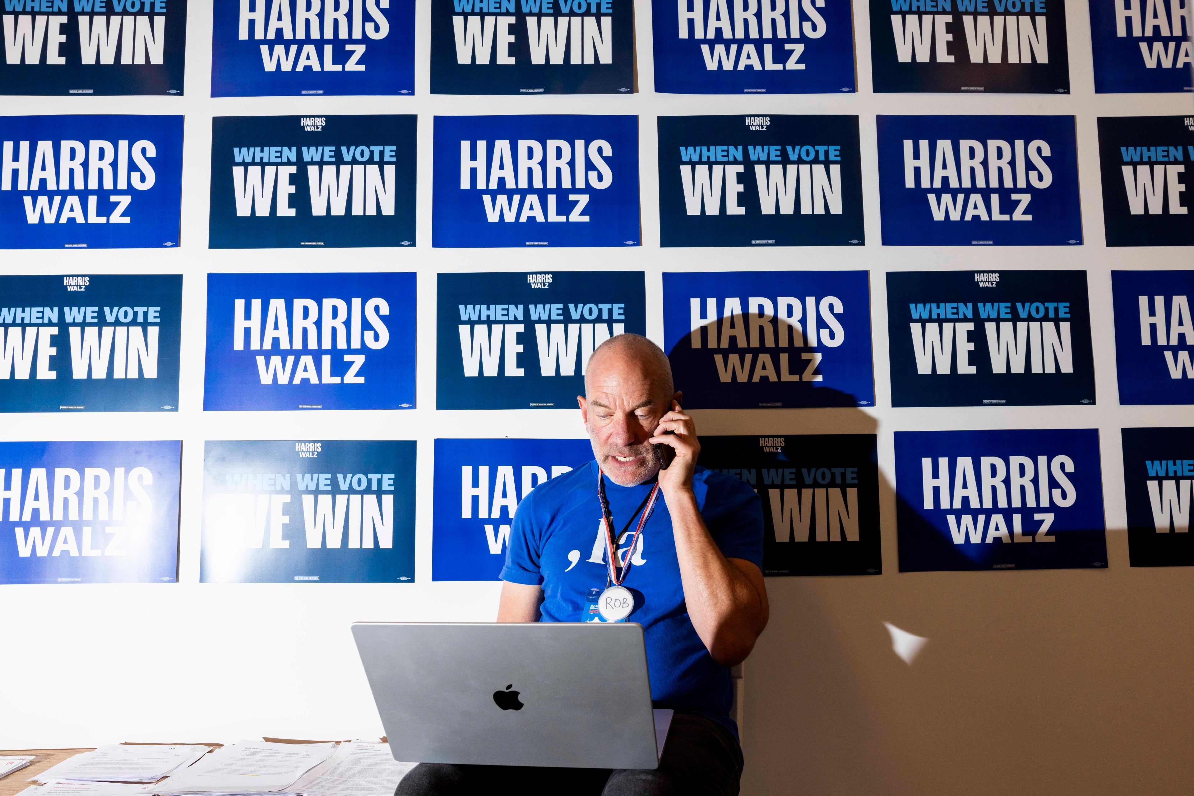 A man sits with a laptop, talking on the phone, in front of a wall covered with blue campaign signs saying &quot;HARRIS WALZ&quot; and &quot;WHEN WE VOTE WE WIN.&quot;