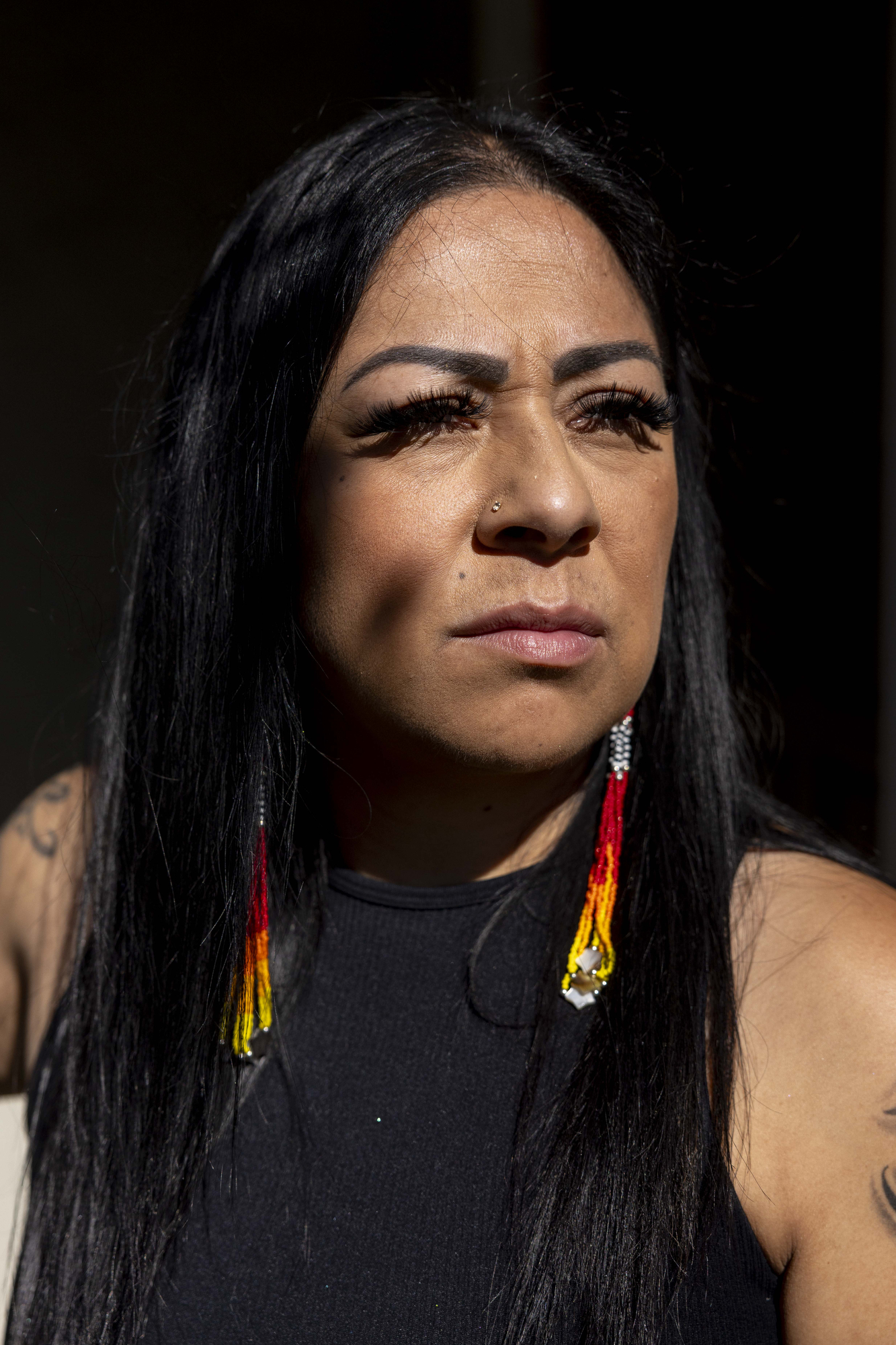 A woman with long black hair and colorful beaded earrings looks pensively into the distance, wearing a black top. Sunlight highlights her facial expression.