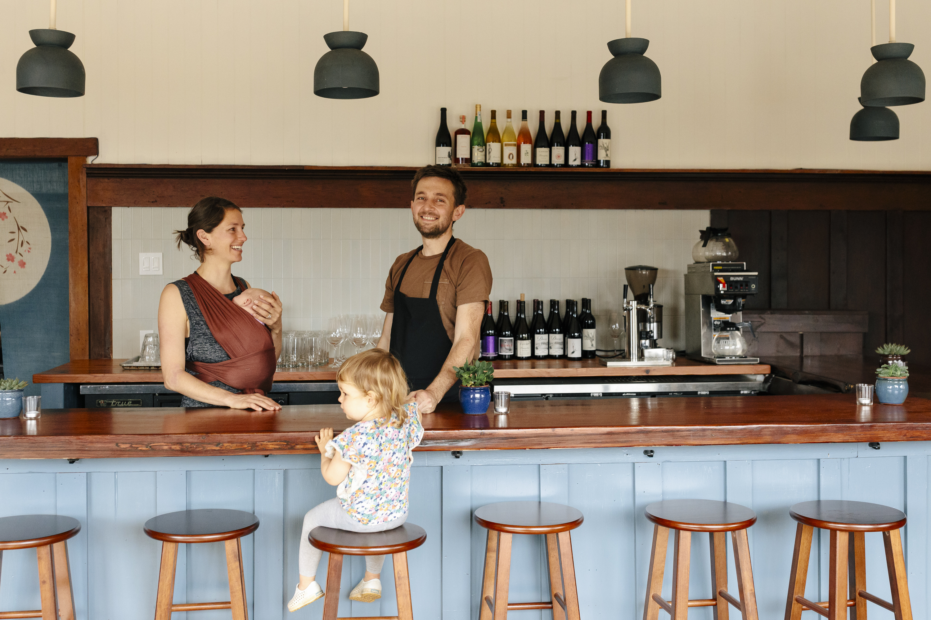 A man and woman smile behind a bar with a small child sitting on a stool. Shelves with bottles and glasses, a coffee machine, and blue pendant lights are visible.