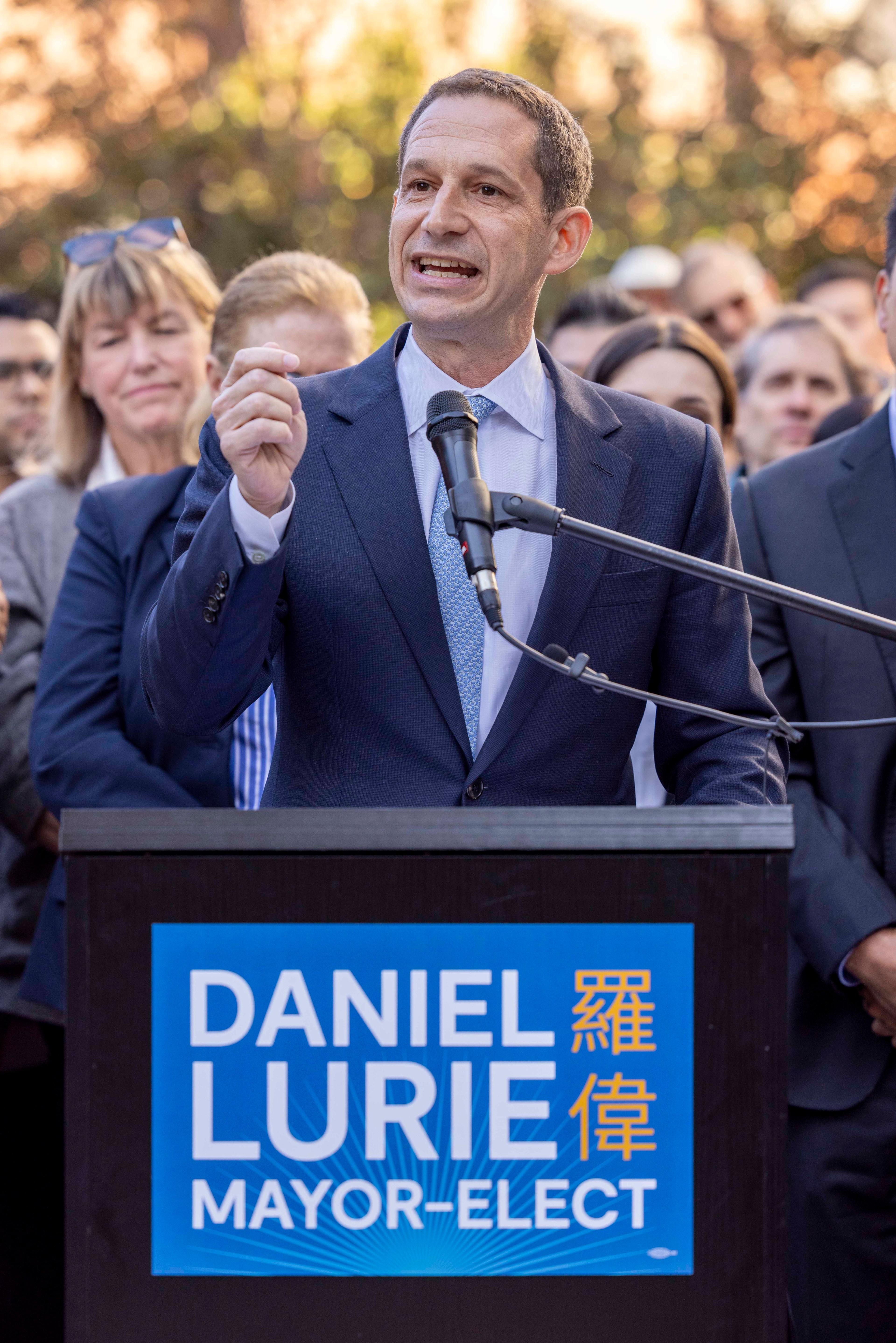 A man in a suit speaks at a podium labeled &quot;Daniel Lurie Mayor-Elect&quot; surrounded by a crowd outdoors, with trees in the background.