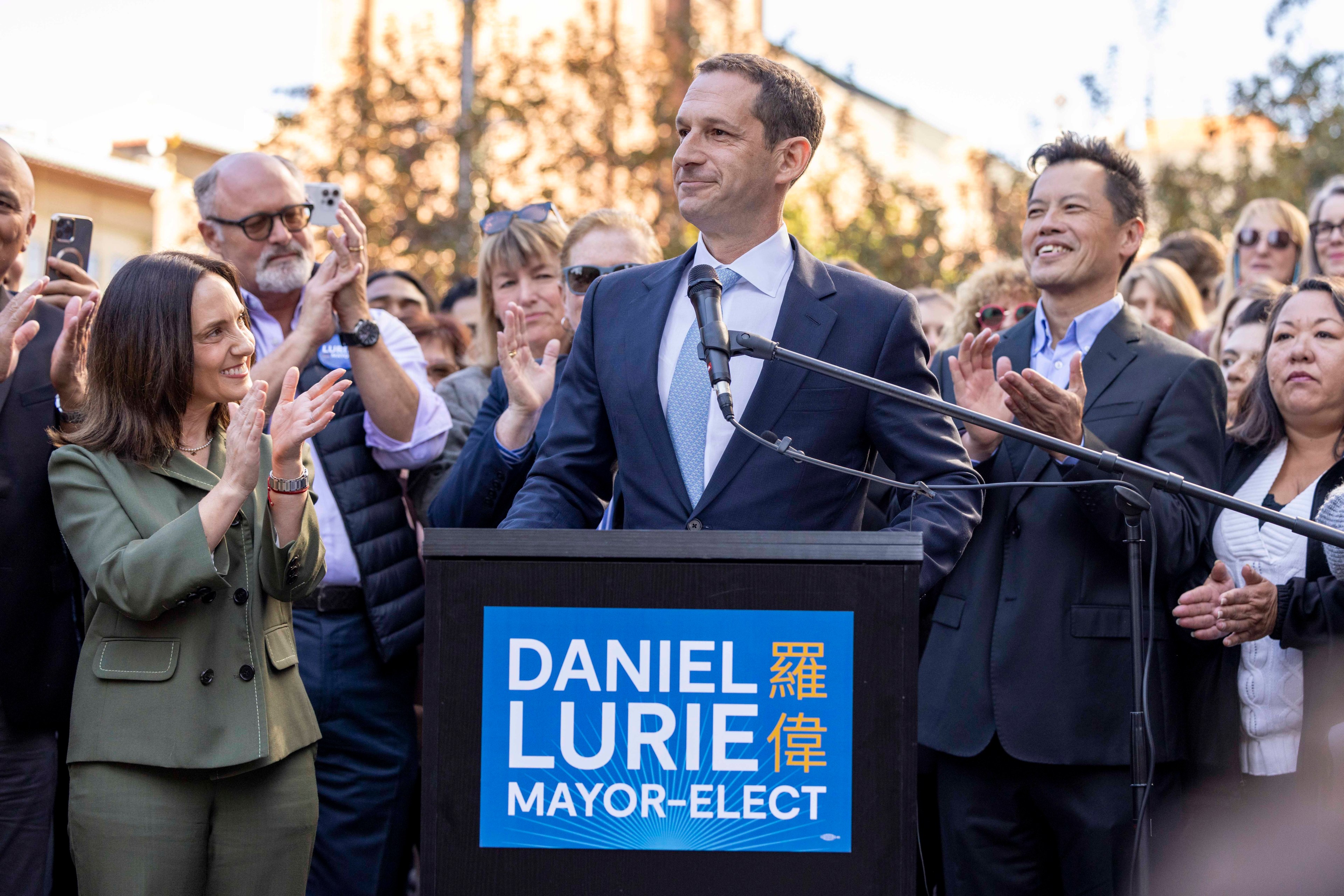 A man stands at a podium labeled "Daniel Lurie Mayor-Elect," smiling, surrounded by a crowd of applauding people in a sunny outdoor setting.