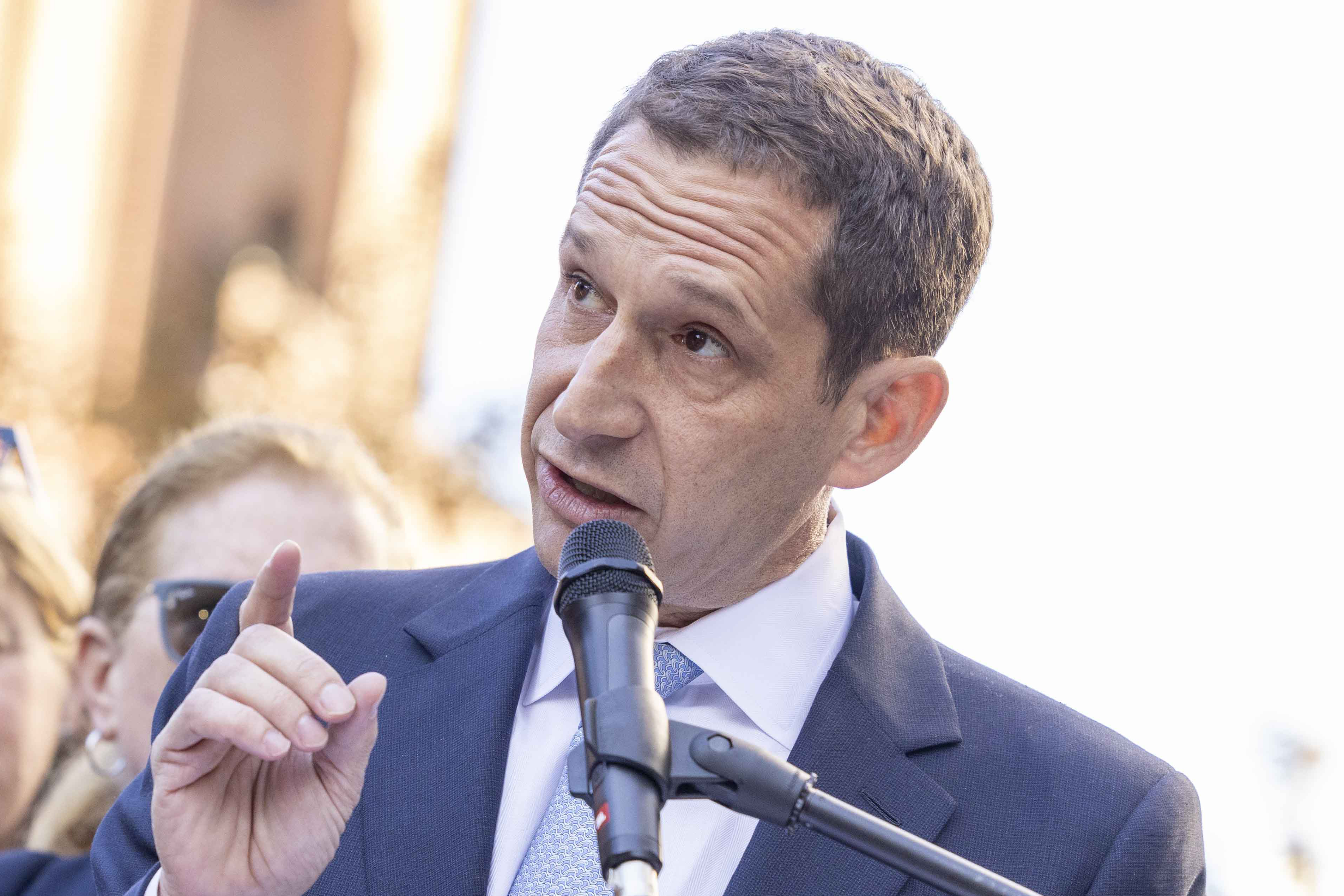 A man in a suit speaks into a microphone, gesturing with one hand. The background is slightly blurred, suggesting an outdoor setting with people.