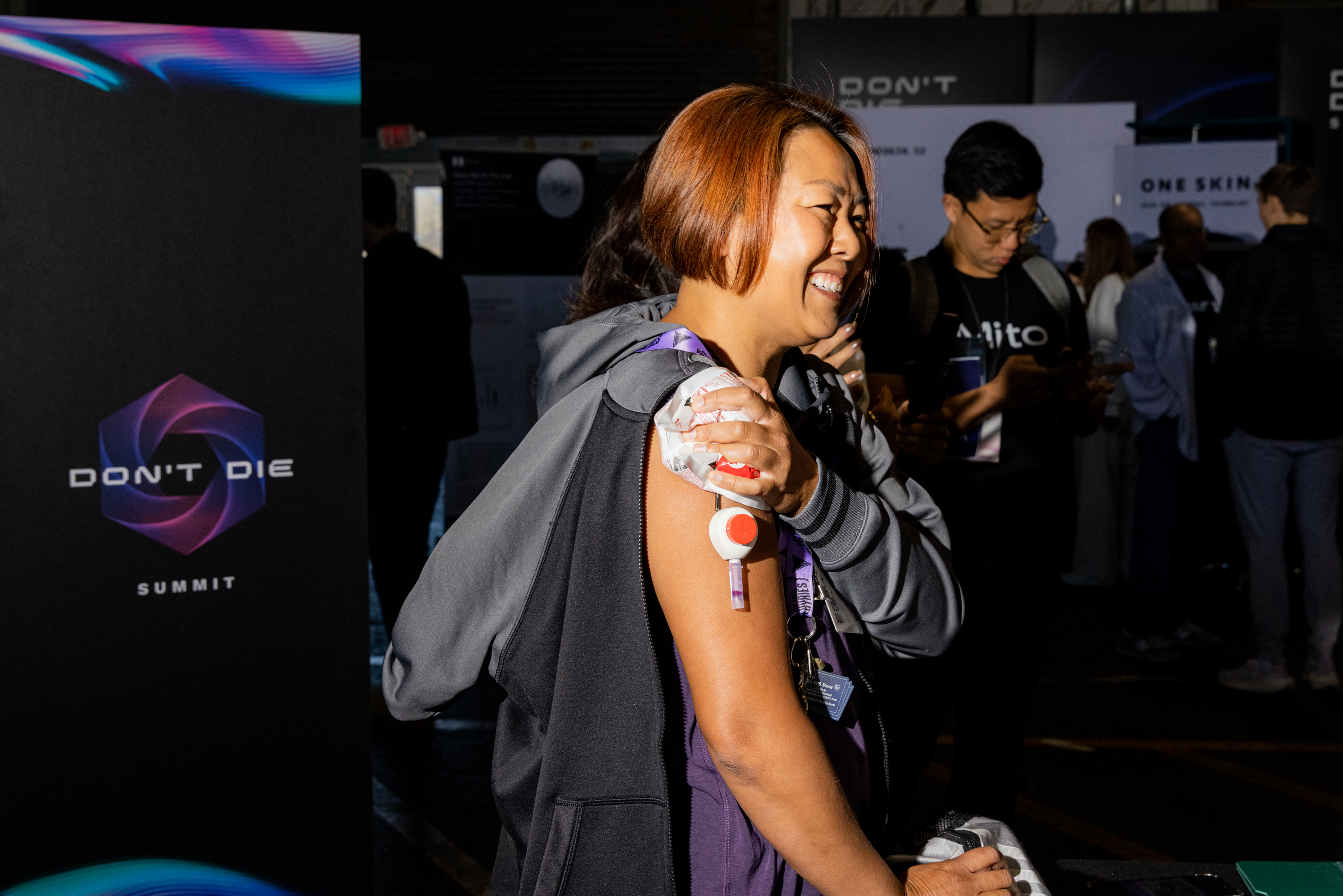 A smiling person in a dark setting is having a medical device attached to their upper arm. There's a &quot;Don't Die Summit&quot; sign in the background.