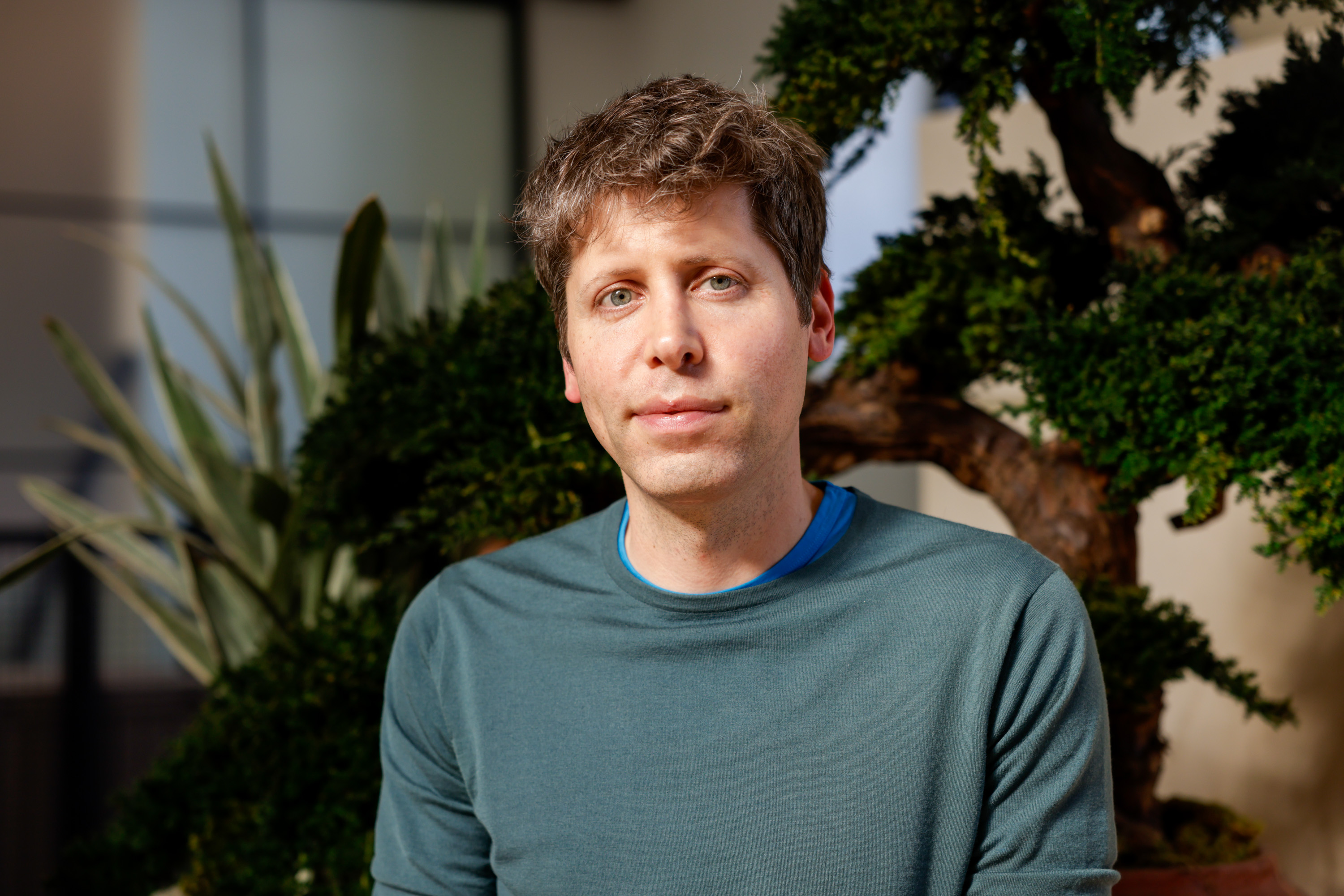 A person with light hair is sitting in front of a lush green plant, wearing a blue shirt under a darker green sweater, against a blurred indoor background.