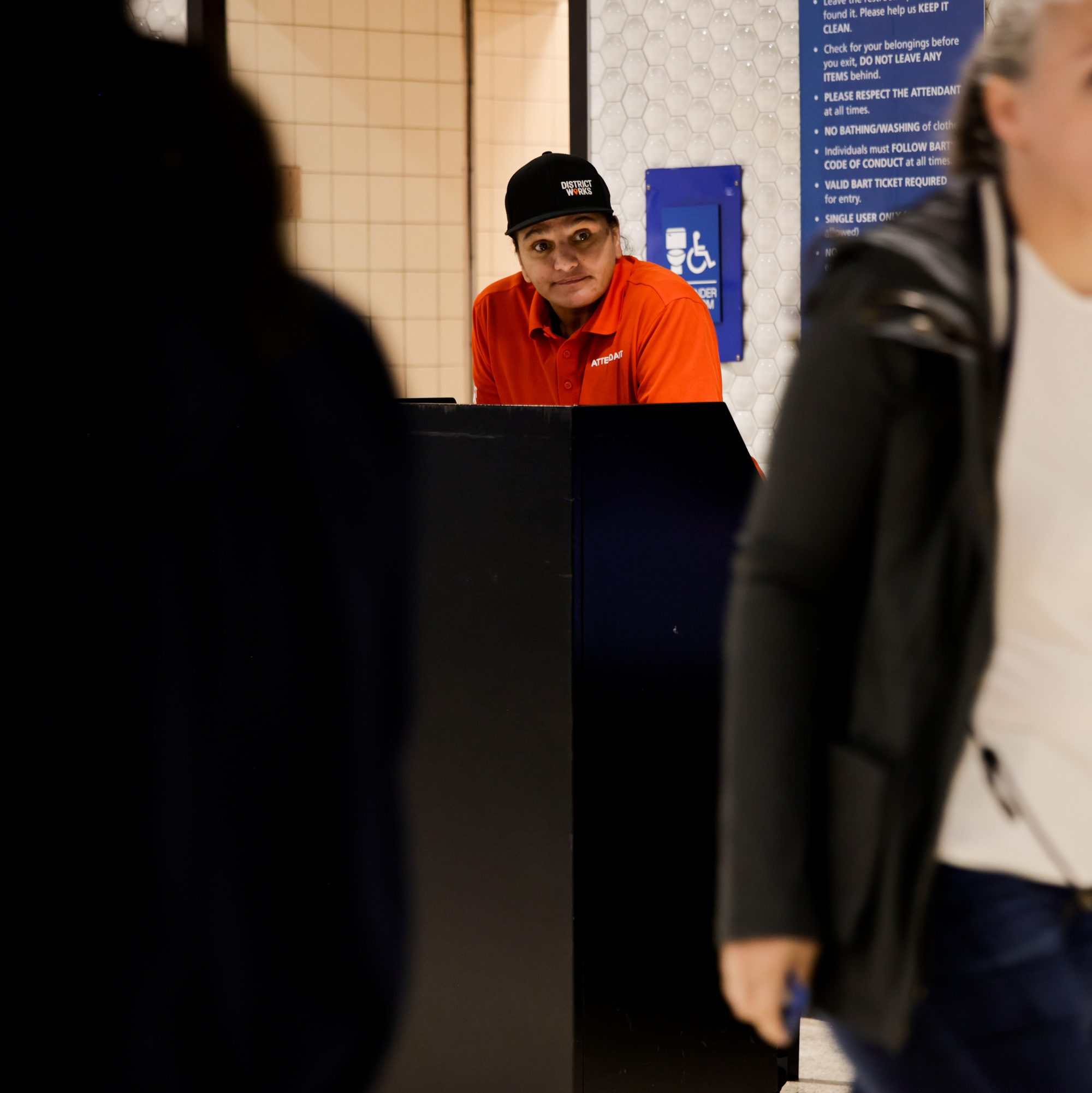 A person in an orange shirt and black cap leans on a counter, with a hexagon-patterned wall and a blue sign behind them. Blurred figures walk past in the foreground.