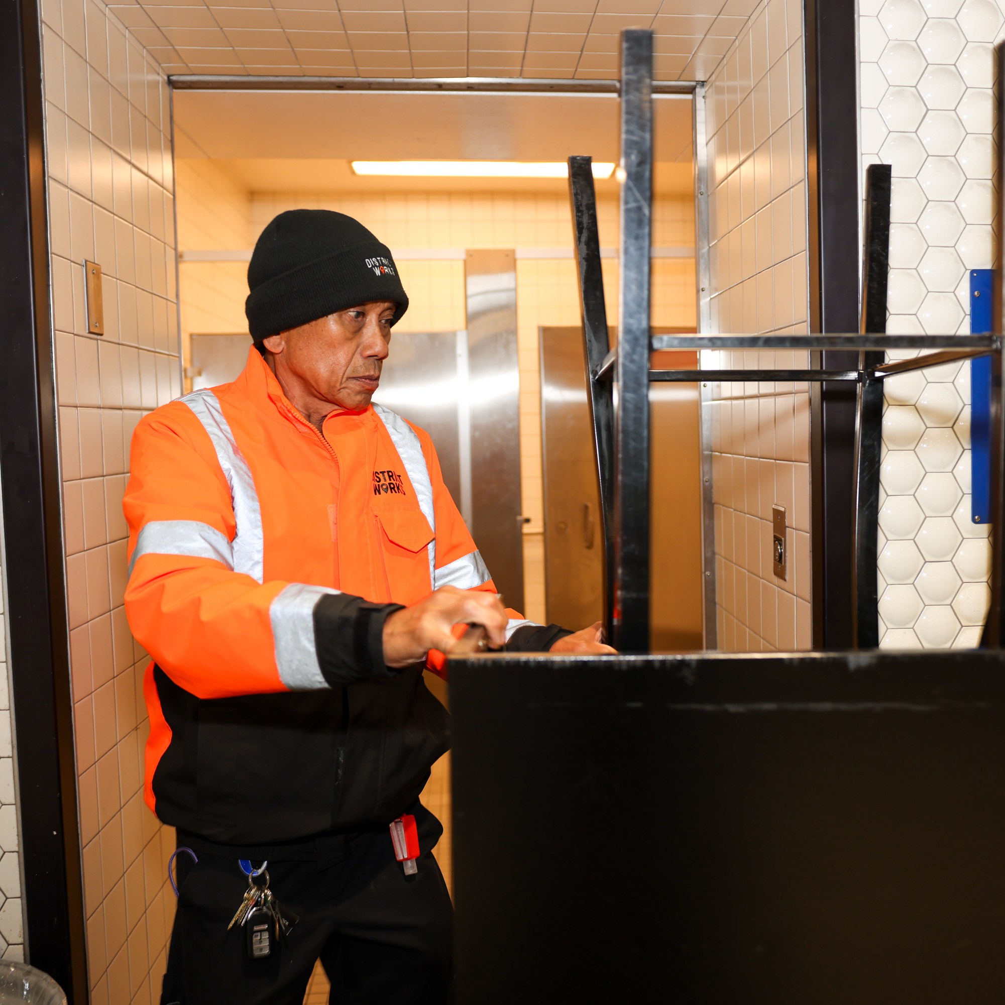 A person in an orange safety jacket and black beanie is in a tiled restroom, cleaning a black object. Signs on the wall indicate it's a multi-gender restroom.