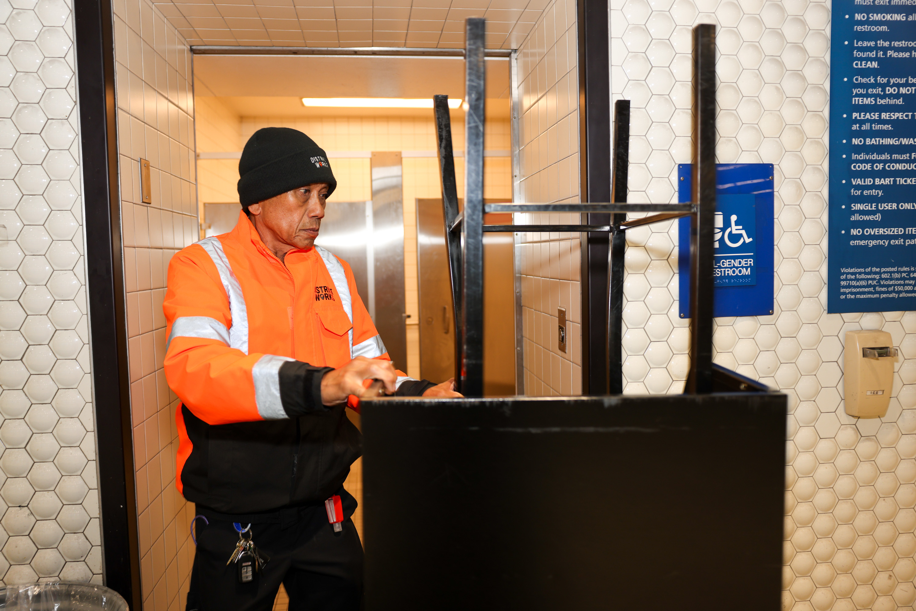 A person in an orange safety jacket and black beanie is in a tiled restroom, cleaning a black object. Signs on the wall indicate it's a multi-gender restroom.