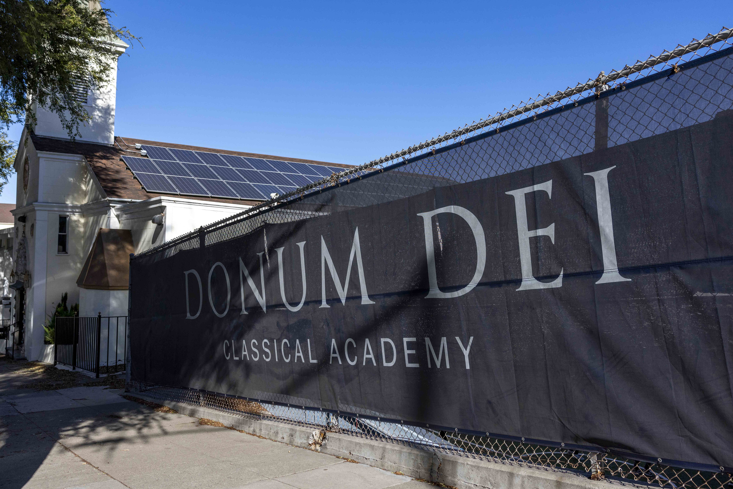 A building with solar panels is visible, partially obscured by a fence with a banner reading &quot;Donum Dei Classical Academy.&quot; The sky is clear and blue.