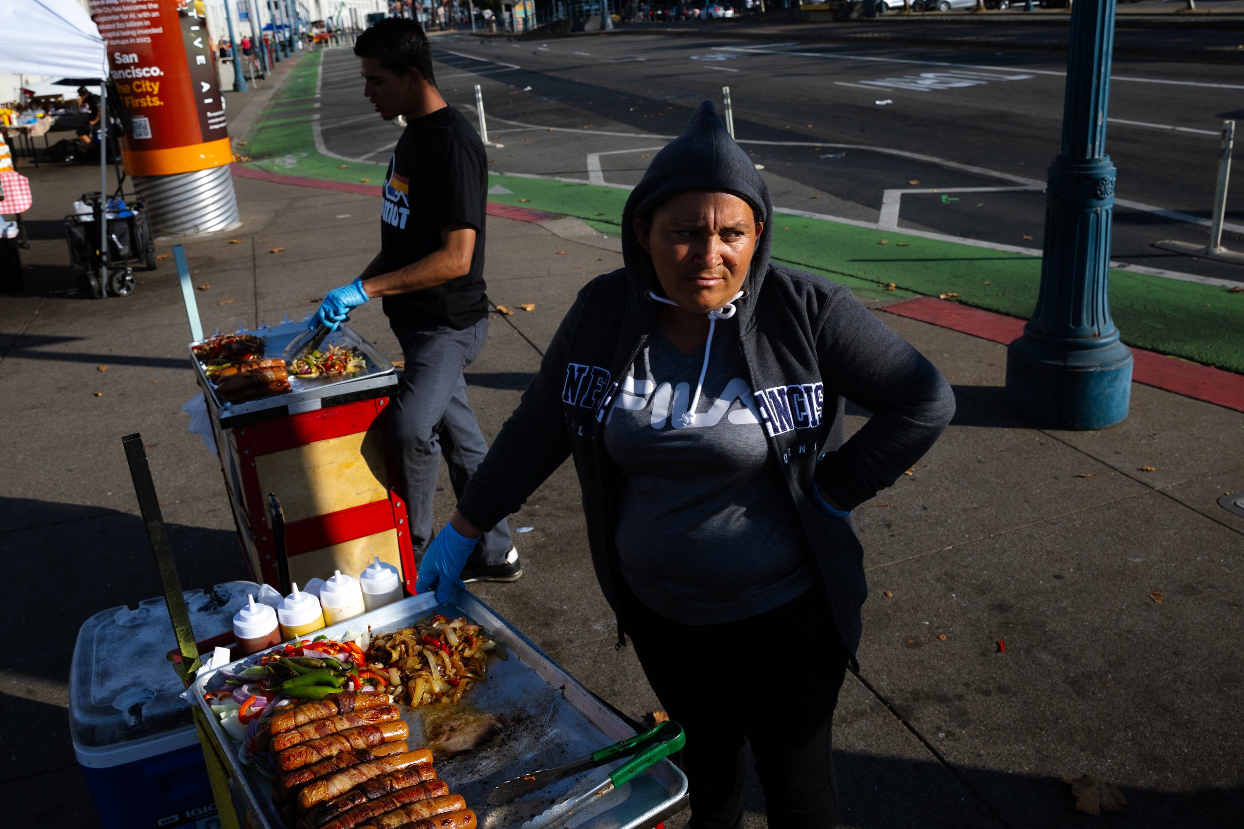 A woman in a hoodie stands next to a food cart with grilled sausages and onions. Another person is preparing food in the background on a city street.