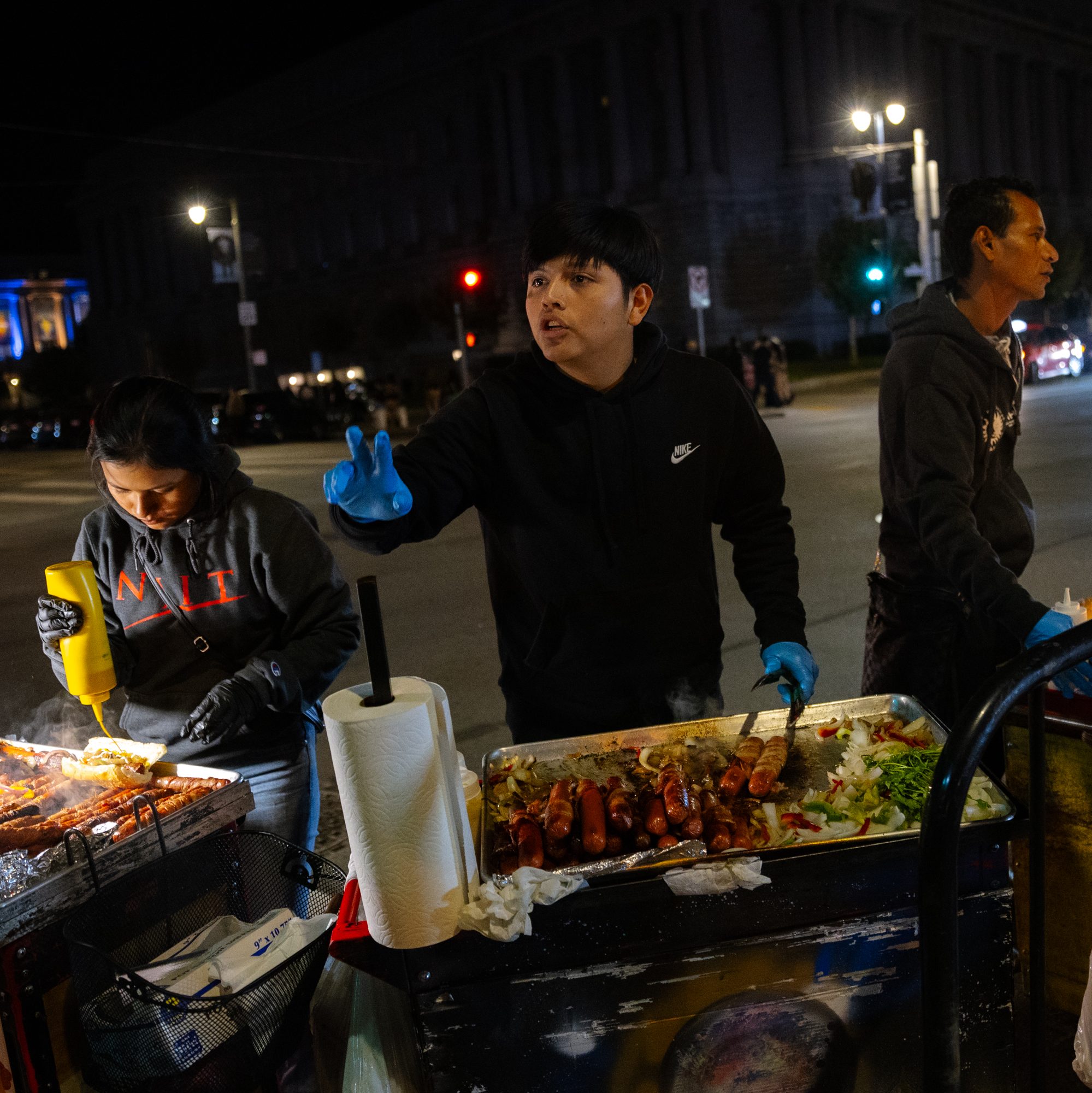 People are working at a nighttime street food stand, grilling sausages and preparing food. One person gestures with a peace sign, and another applies mustard.