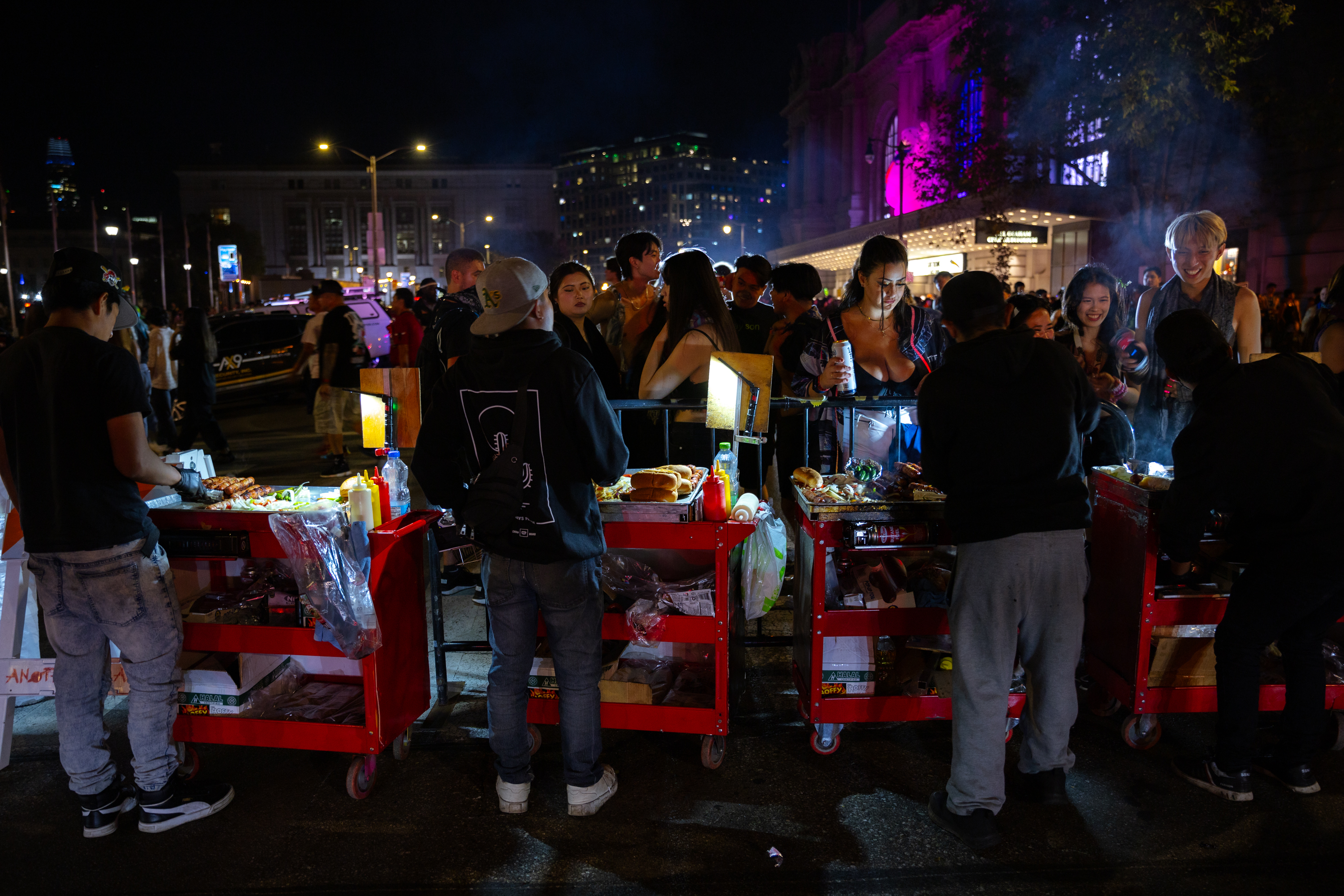 A bustling street scene shows people gathering around several illuminated food carts at night, with a vibrant city background and colorful lights illuminating the area.