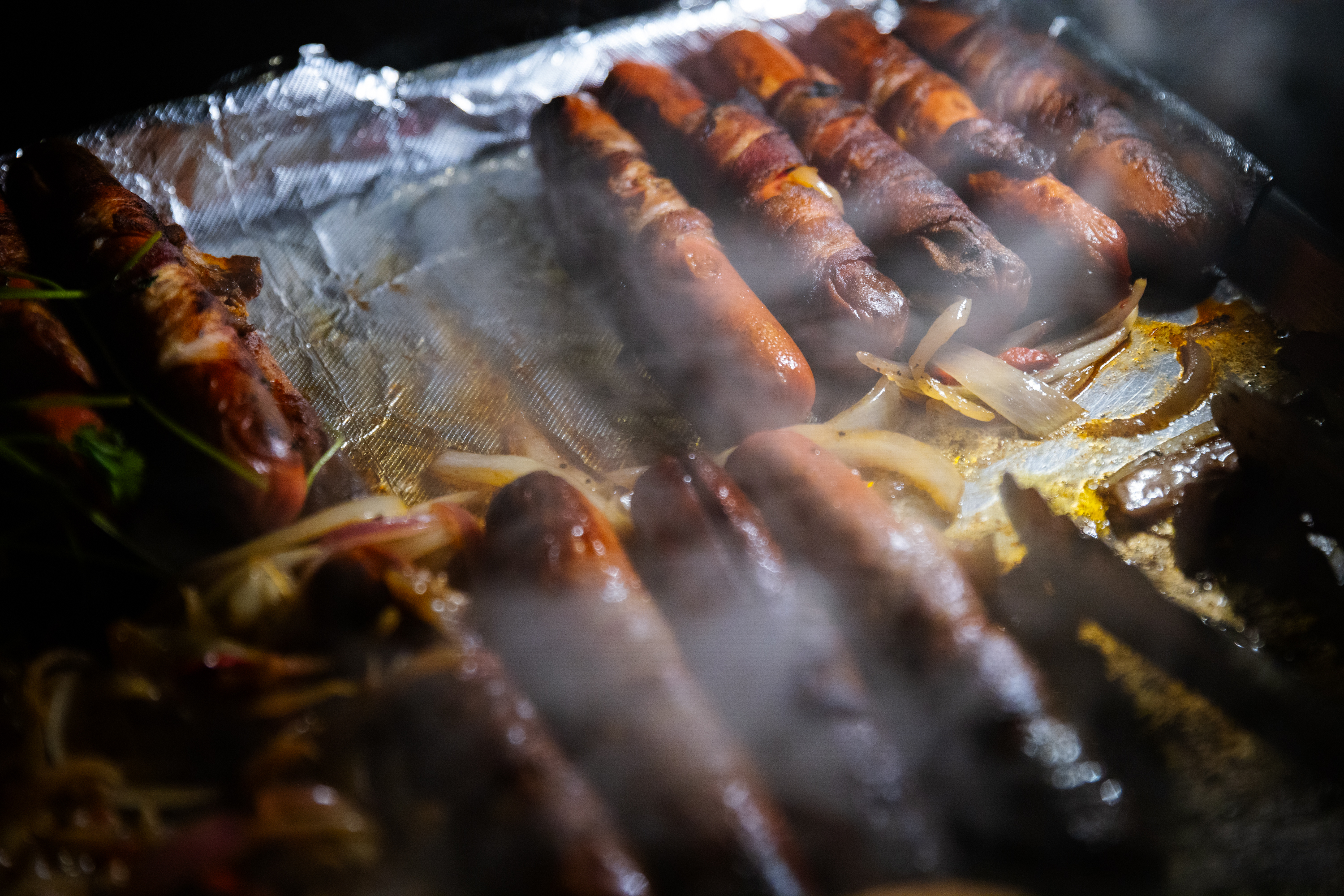 The image shows several sausages wrapped in bacon grilling, with onions scattered on a foil-covered surface. Steam rises, creating a smoky atmosphere.