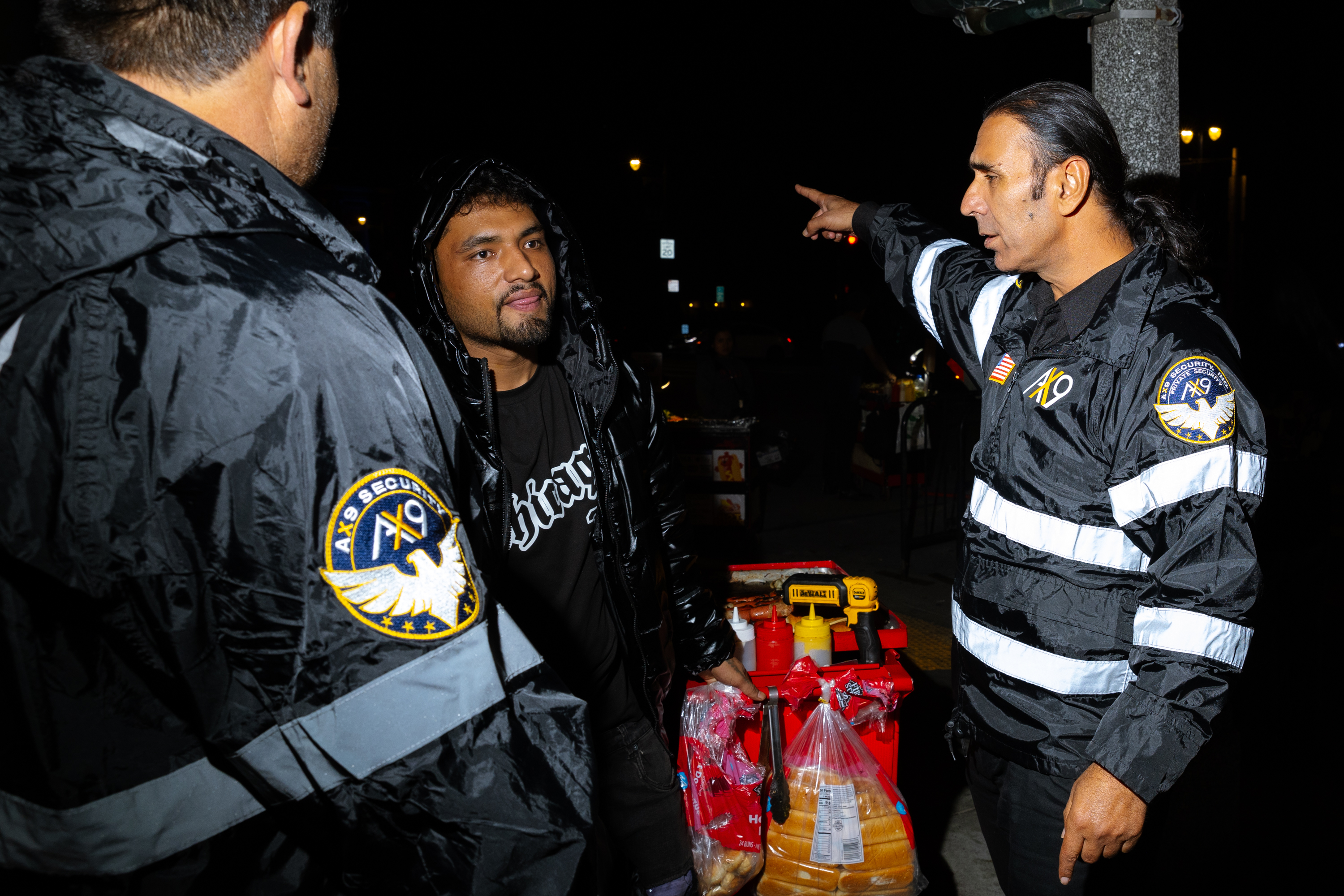 Two security personnel in reflective jackets are interacting with a man in a black hoodie. One officer is pointing while standing near a cart with food items.