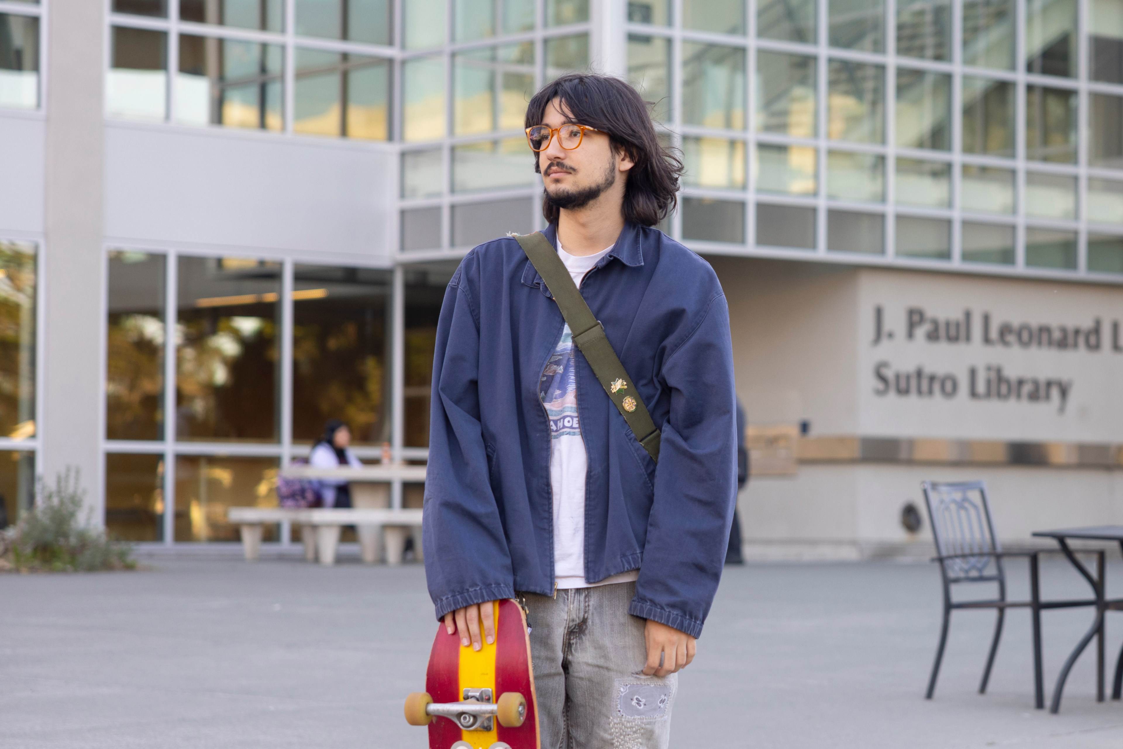 A person with glasses and long hair is holding a red skateboard outdoors. They wear a blue jacket and a green strap. The library building is in the background.