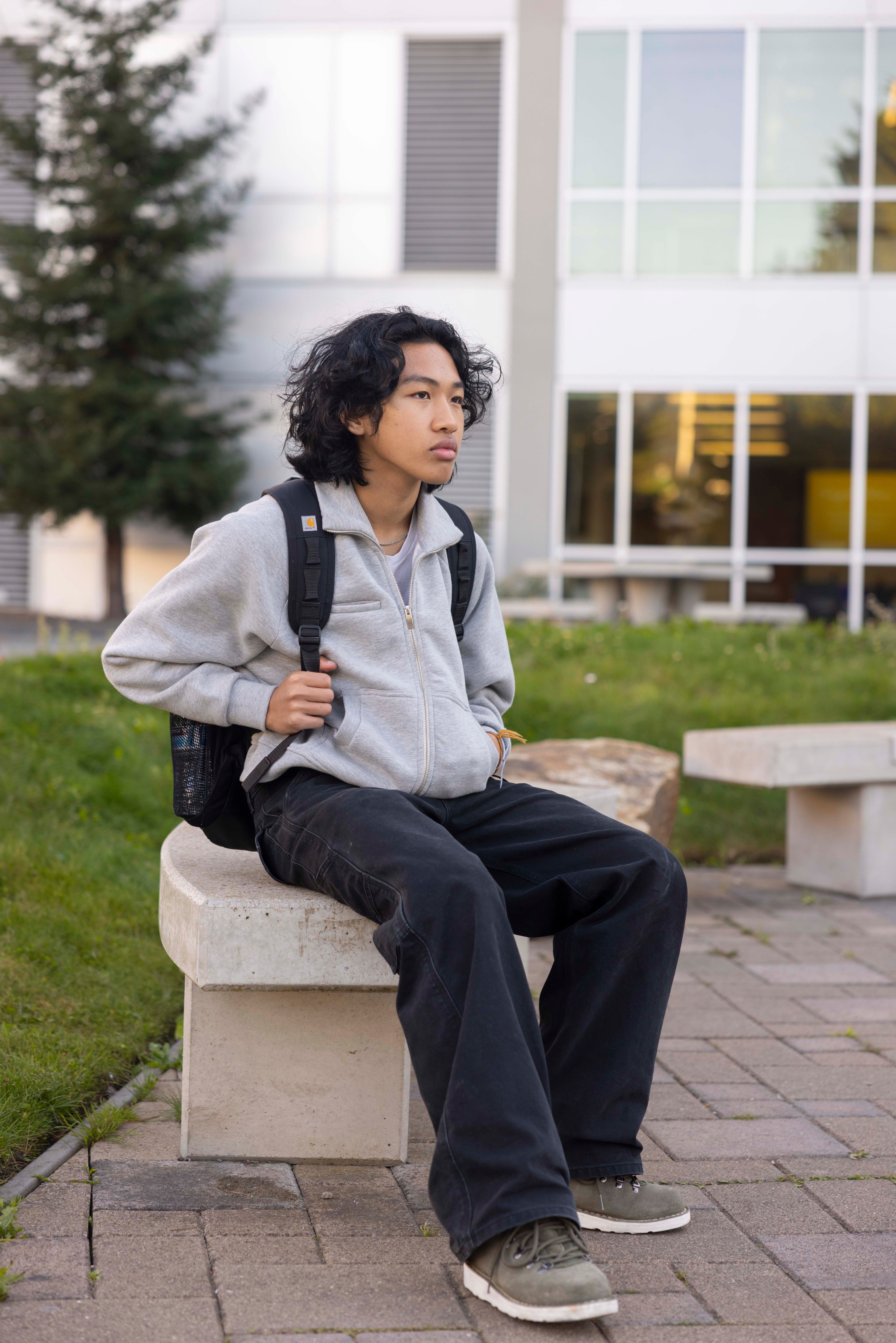 A person with wavy hair sits on a concrete bench, wearing a gray jacket and black pants, with a backpack. The background shows a building and greenery.