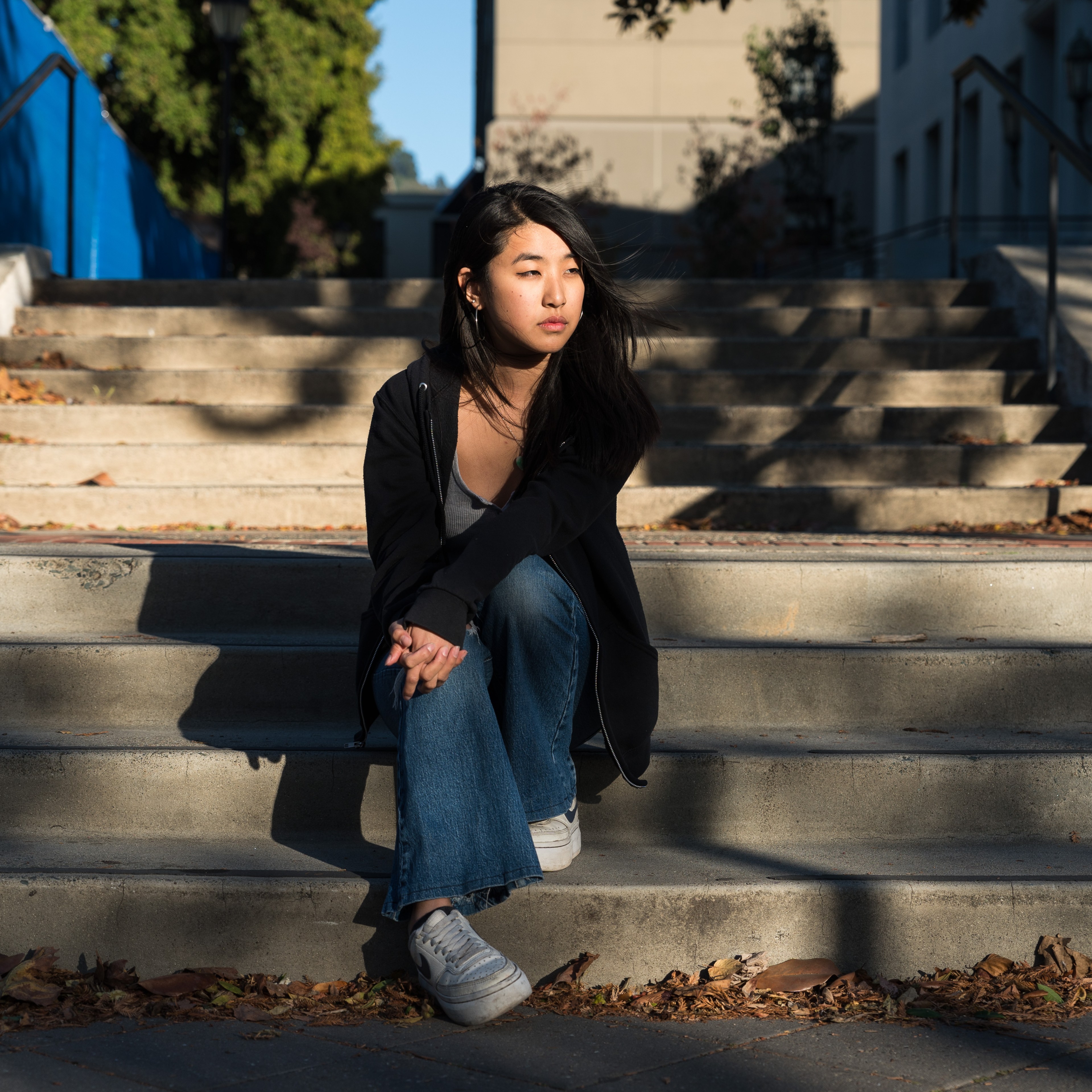 A person is sitting on outdoor steps, wearing a black jacket and jeans, with sunlight casting shadows. The background includes trees and a blue wall.