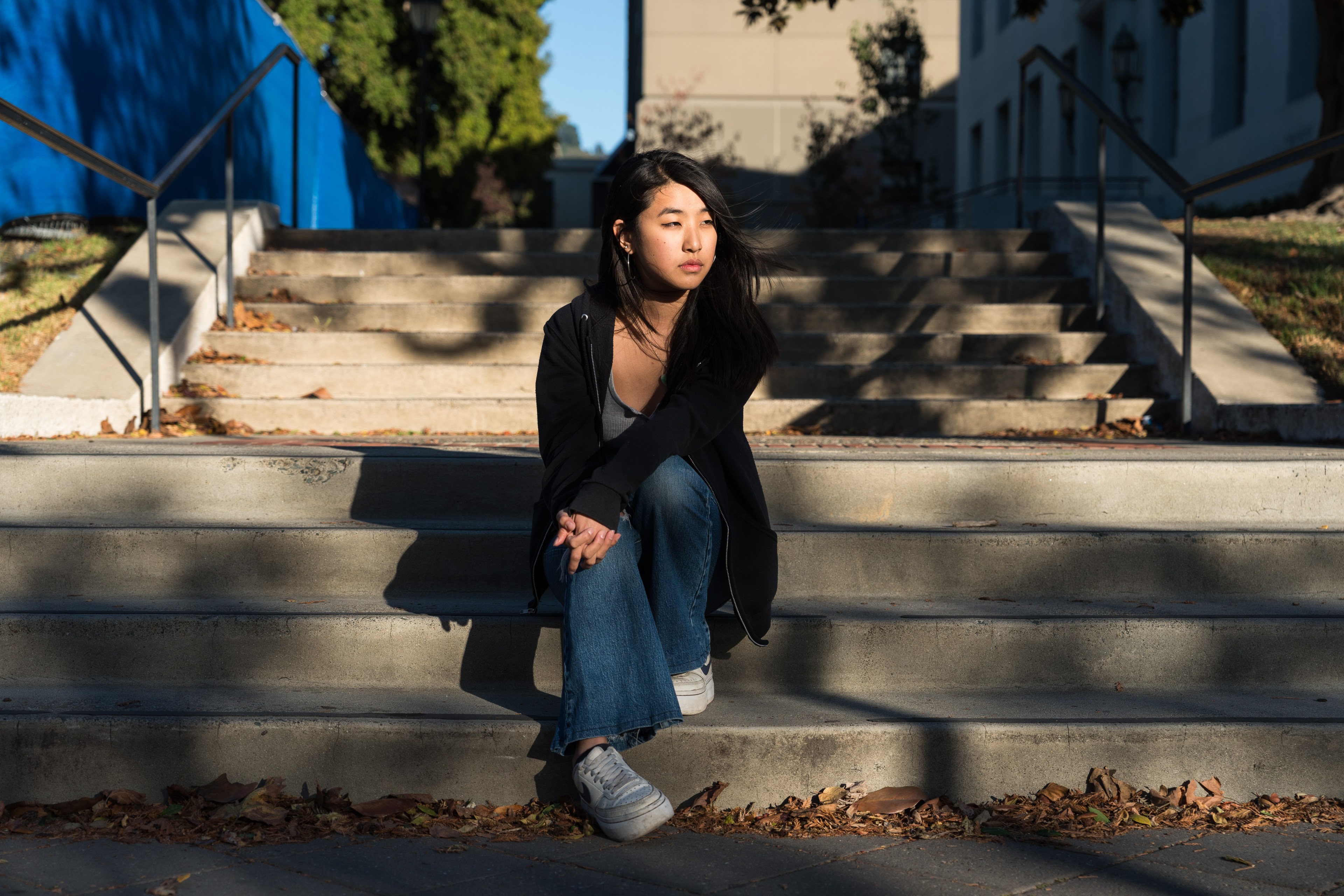 A person is sitting on outdoor steps, wearing a black jacket and jeans, with sunlight casting shadows. The background includes trees and a blue wall.