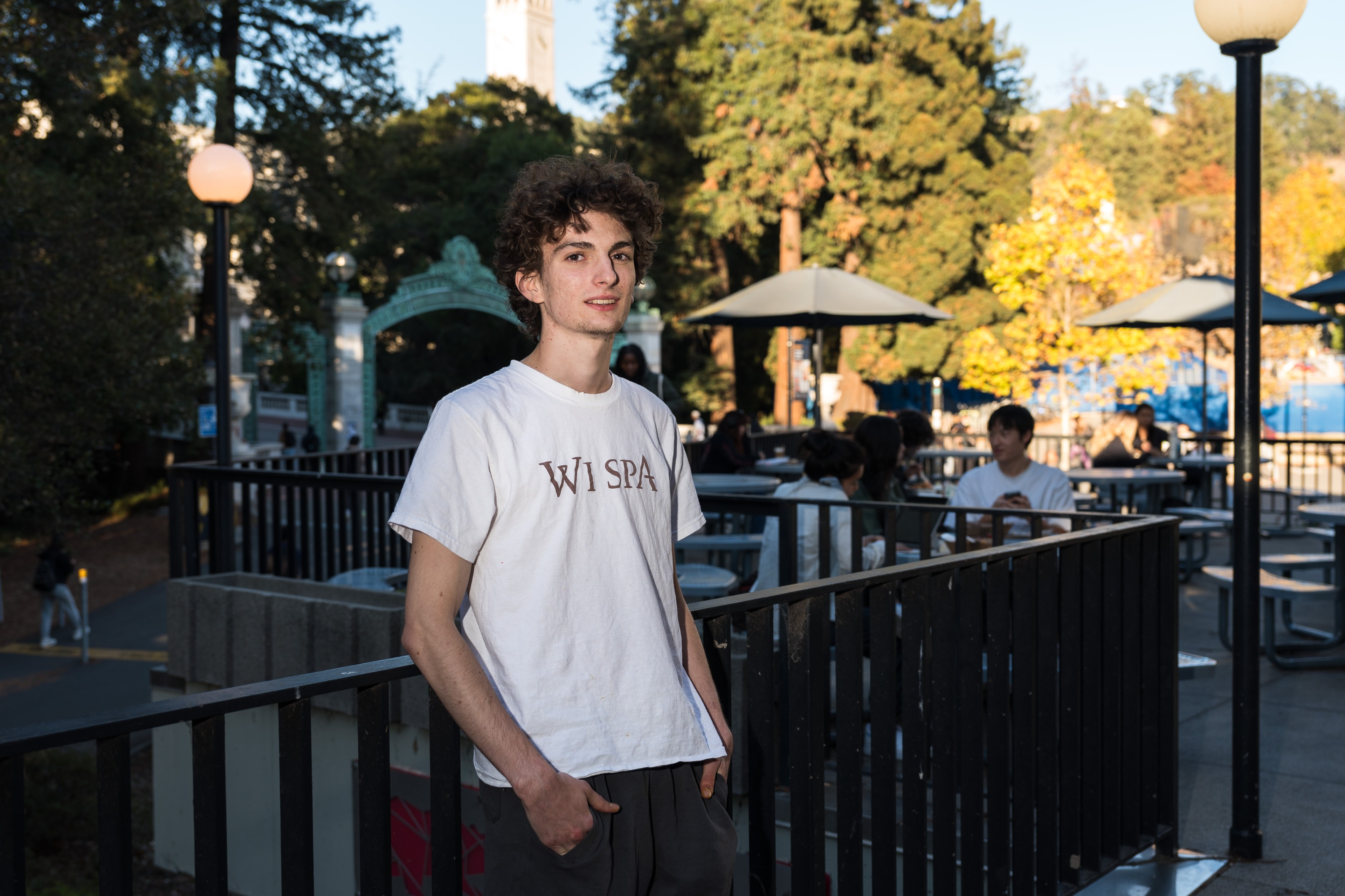 A young man with curly hair stands outdoors, wearing a white shirt with &quot;Wi Spa&quot; printed on it. Behind him are trees, people sitting at tables, and lamp posts.