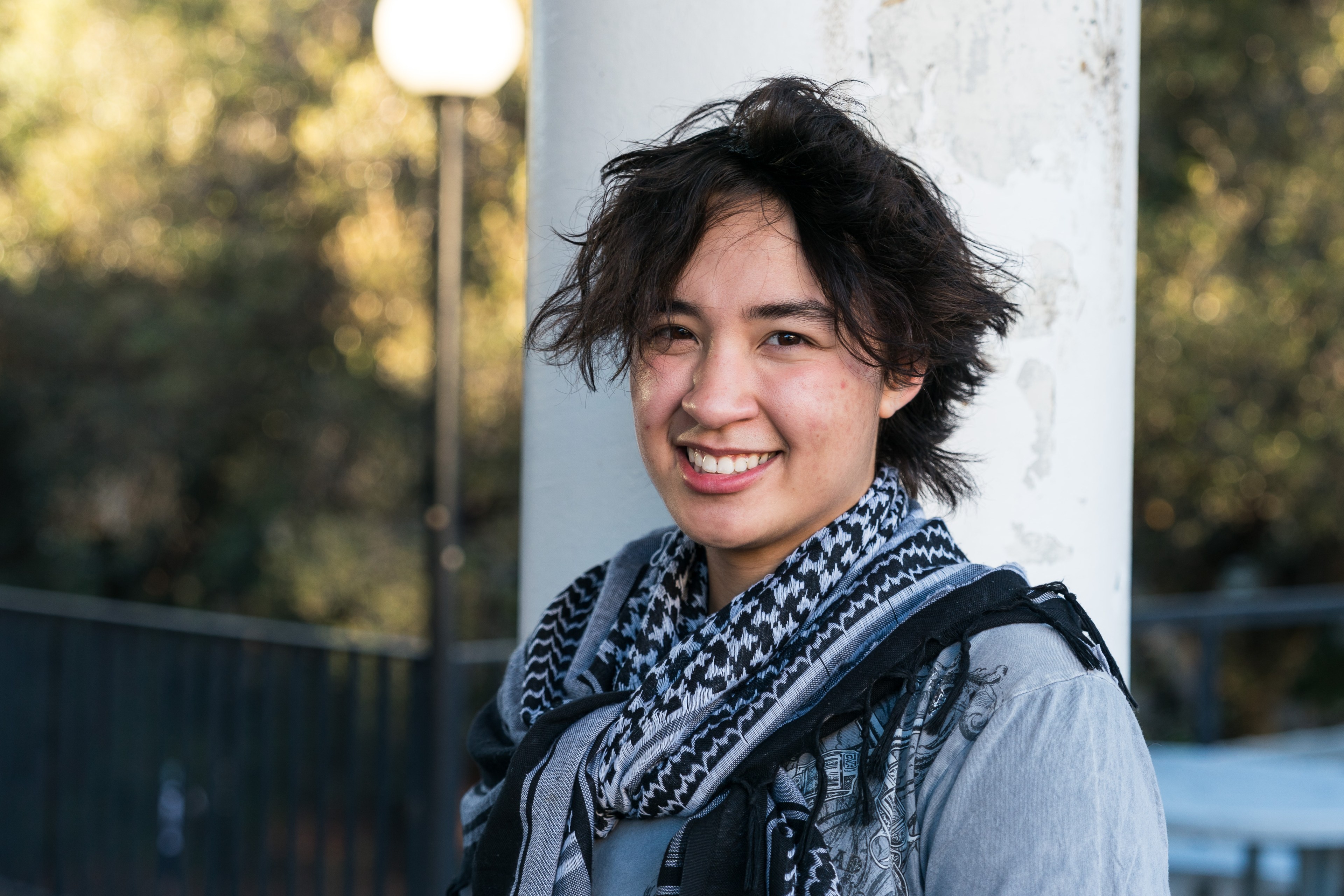 A person with short, tousled hair smiles warmly, wearing a patterned scarf and jacket. They stand outside against a pillar, with trees and a lamppost in the background.