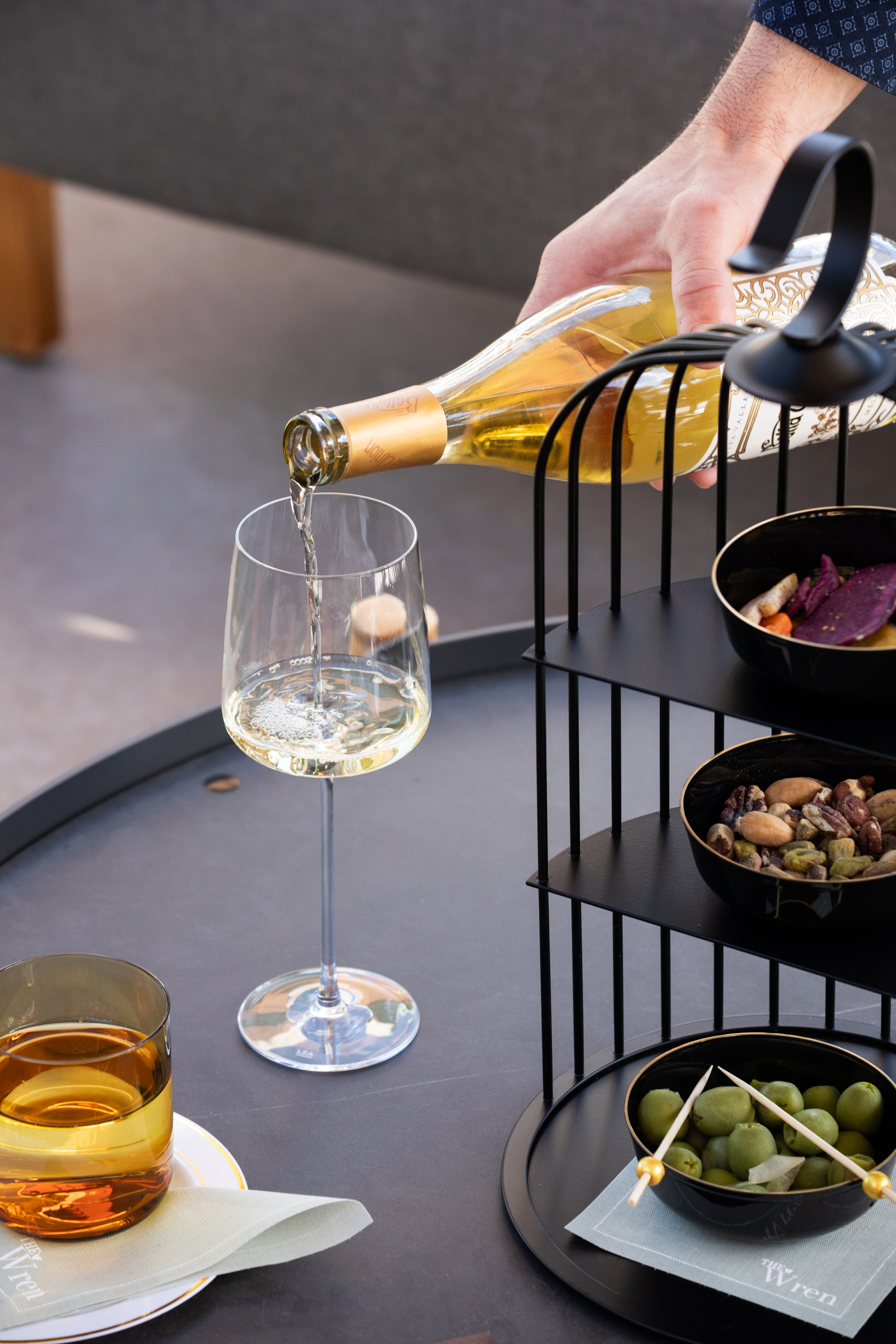 A hand pours white wine into a glass on a table. Nearby is another drink in a glass, and a tiered tray holds bowls of nuts, olives, and chips.