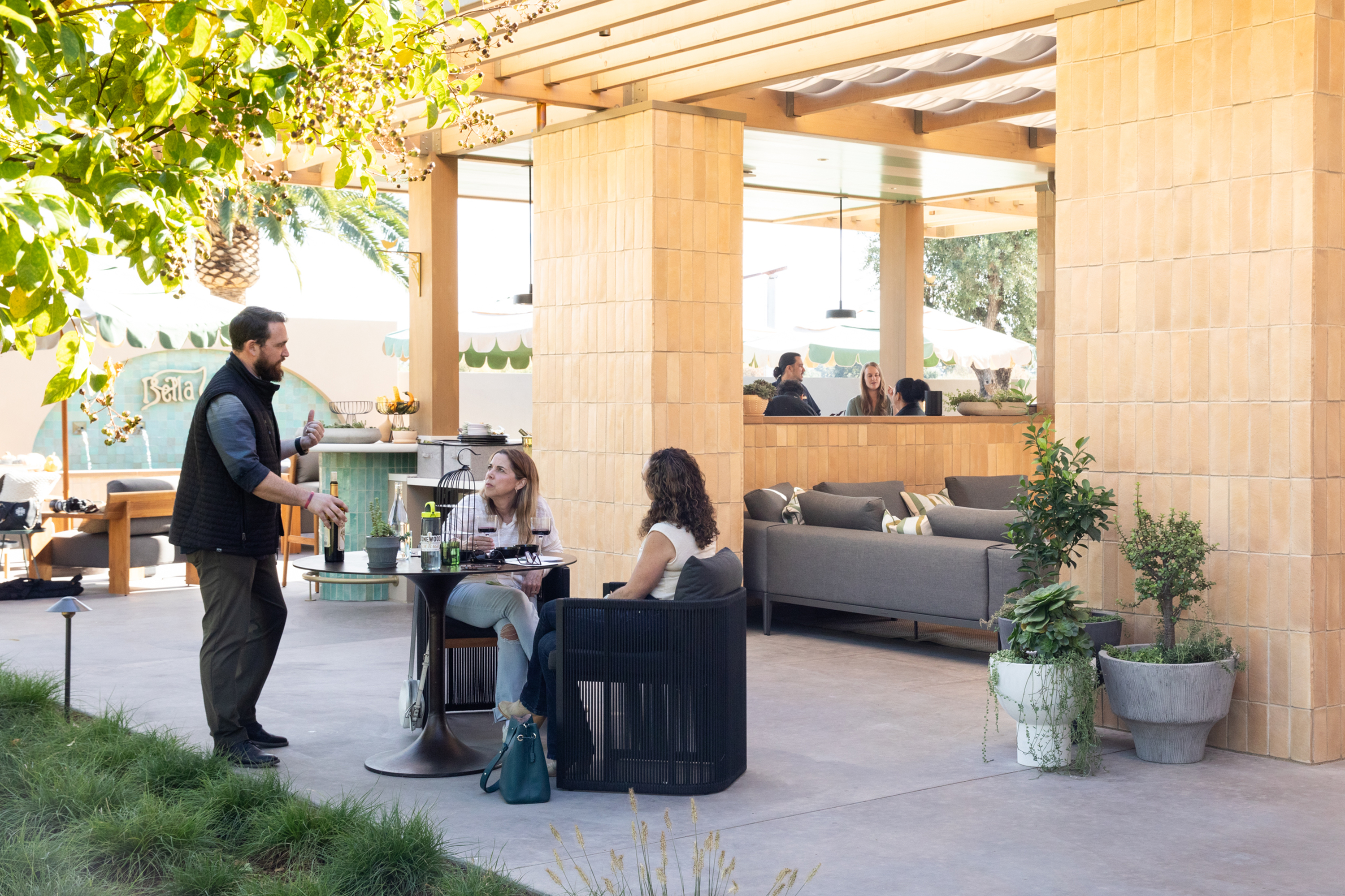 A waiter serves two women at an outdoor cafe with wooden structures. They're seated at a small table under leafy branches, surrounded by plants and plush seating.