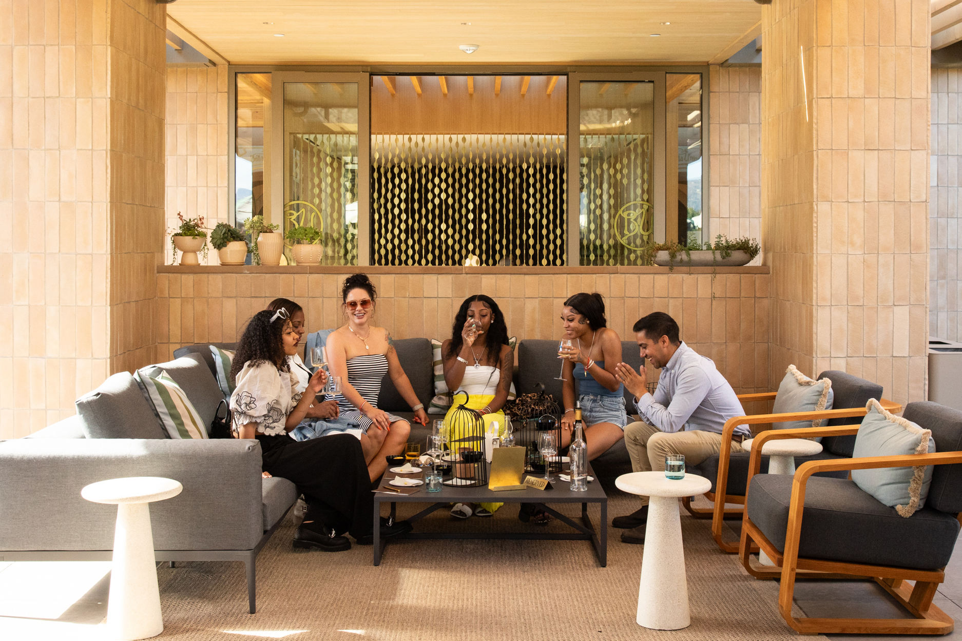 A group of five people sit in a stylish outdoor lounge area, enjoying drinks and chatting. The space is cozy with modern furniture and potted plants.