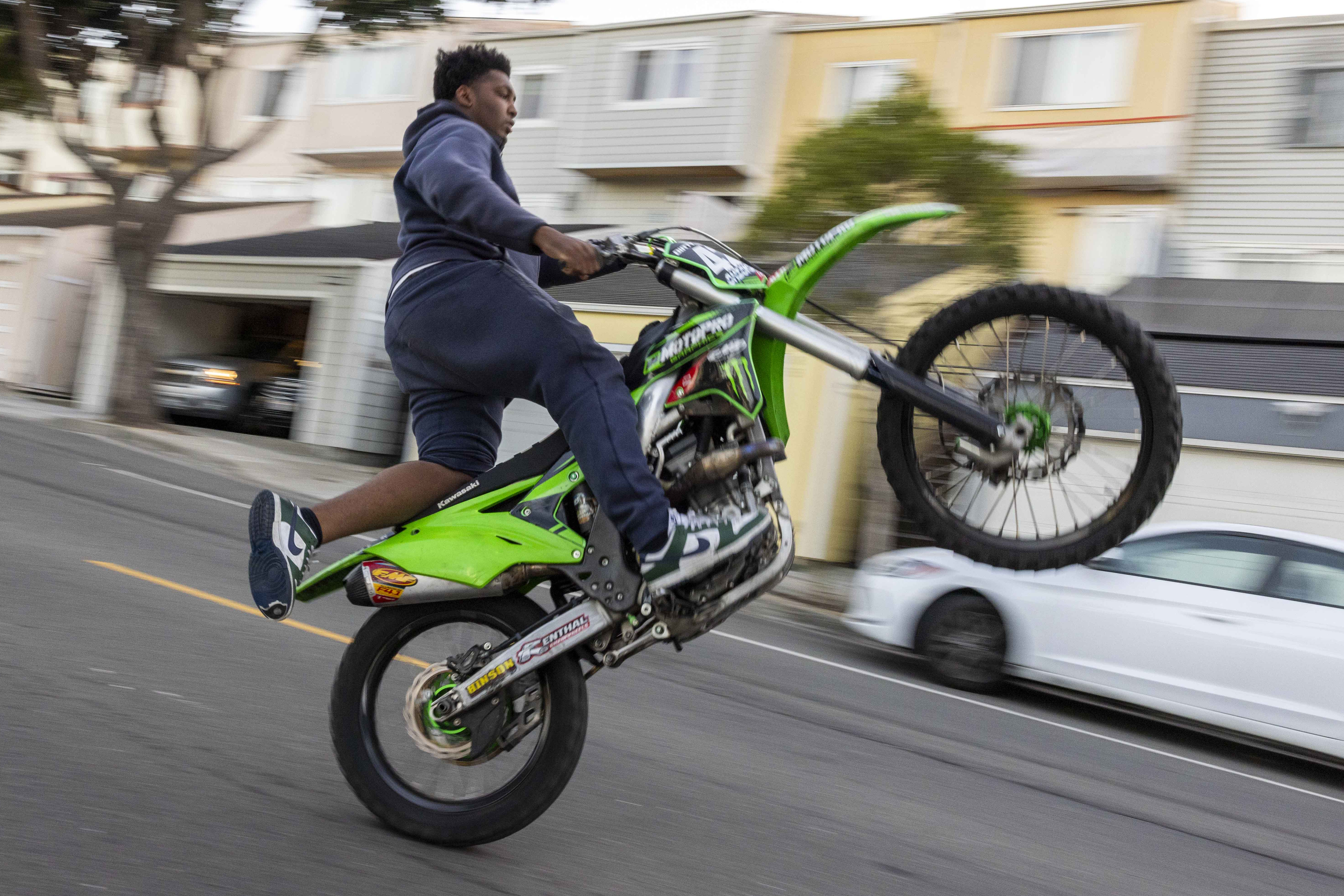 A person performs a wheelie on a green motorcycle in a residential street, with houses blurred in the background, capturing motion and excitement.