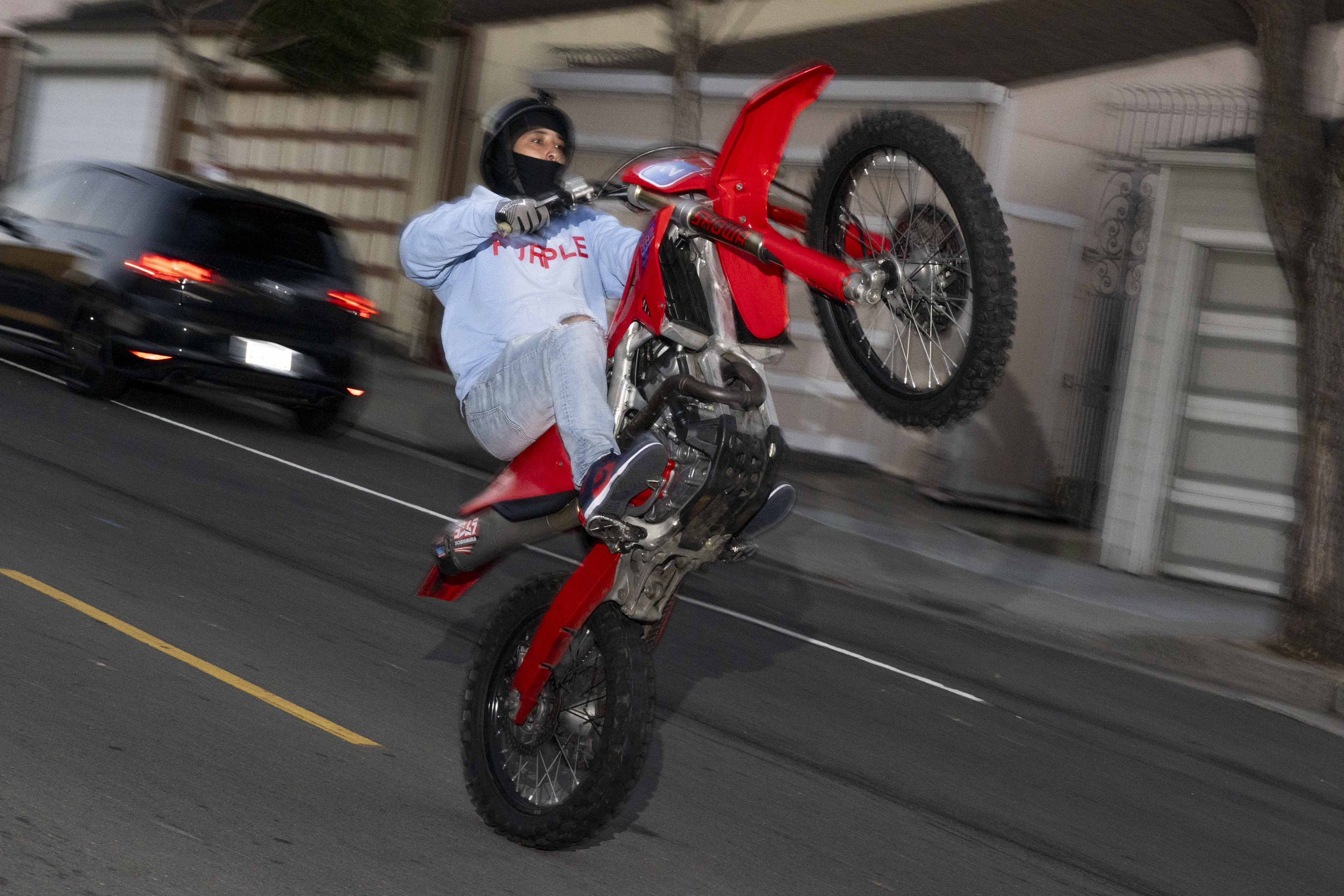 A person in a helmet does a wheelie on a red dirt bike down a city street, wearing a light blue hoodie and jeans, while cars and houses blur in the background.