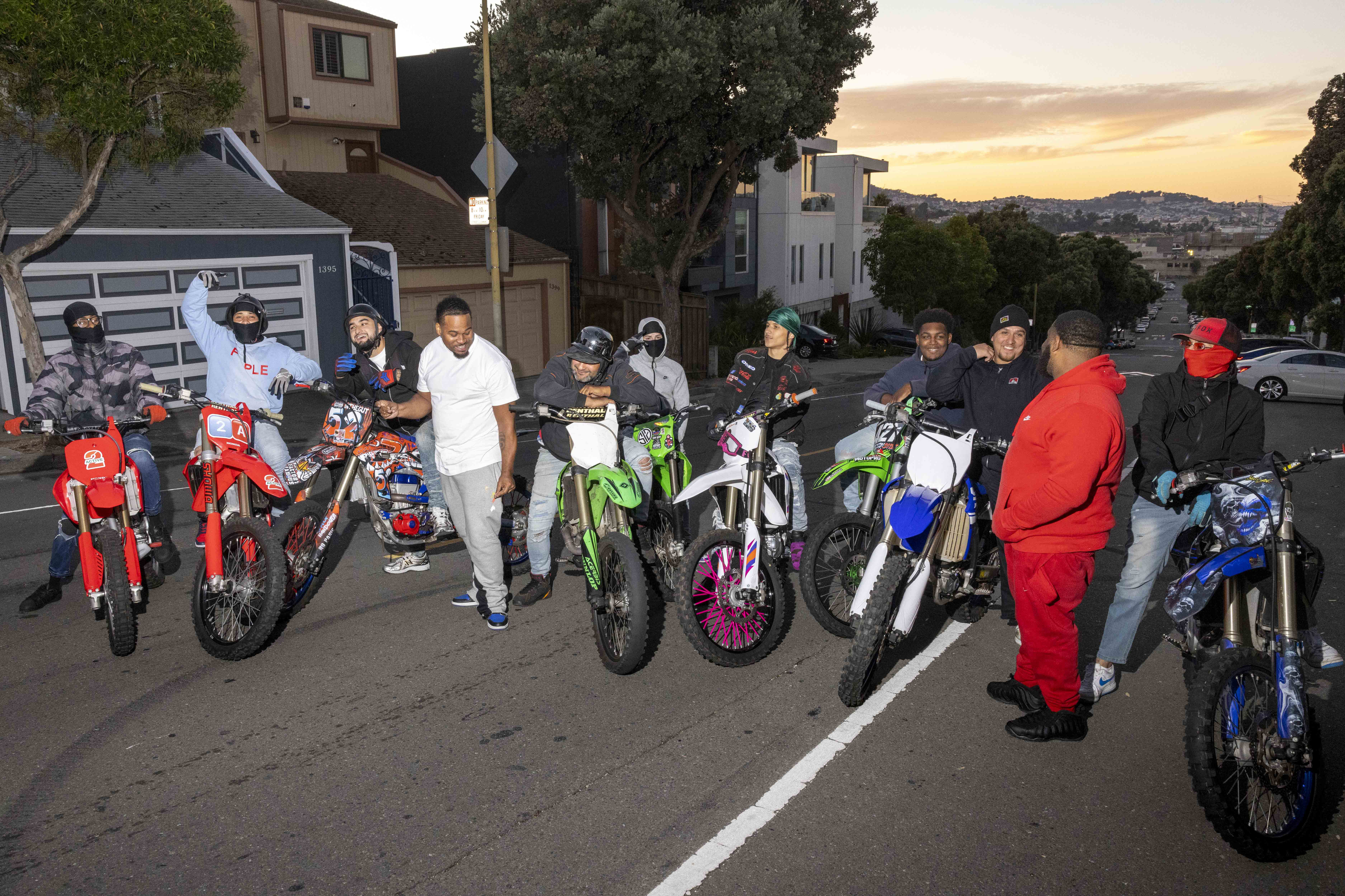 A group of people with dirt bikes gathers on a suburban street at sunset. They appear relaxed and cheerful, with some standing and others sitting on their bikes.