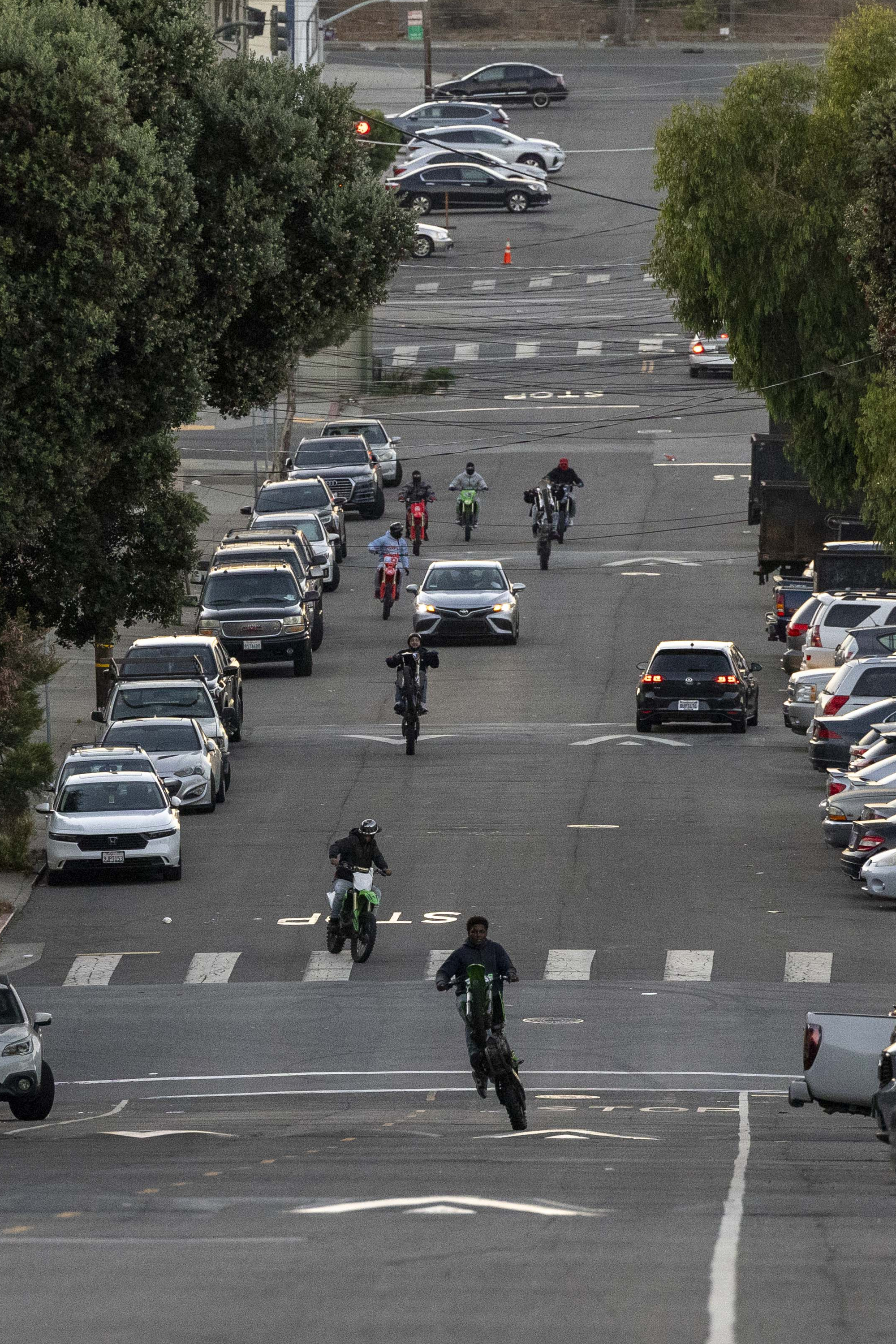 A group of motorcyclists rides up a sloping urban street, some performing wheelies. Cars are parked on both sides, and trees line the sidewalks.