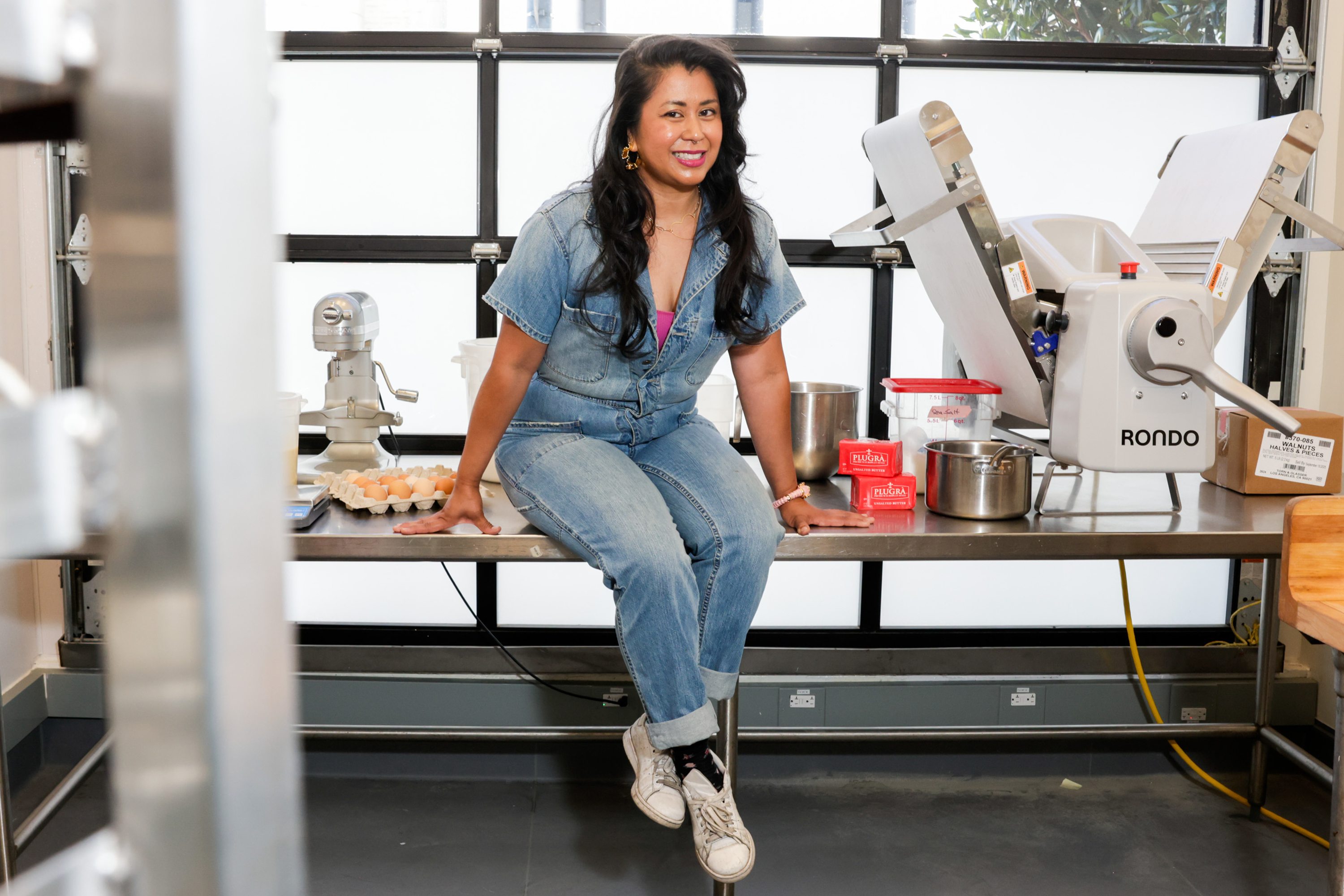 A woman in denim sits on a kitchen counter, surrounded by baking supplies like eggs, flour, and a mixer, with a dough sheeter in the background.