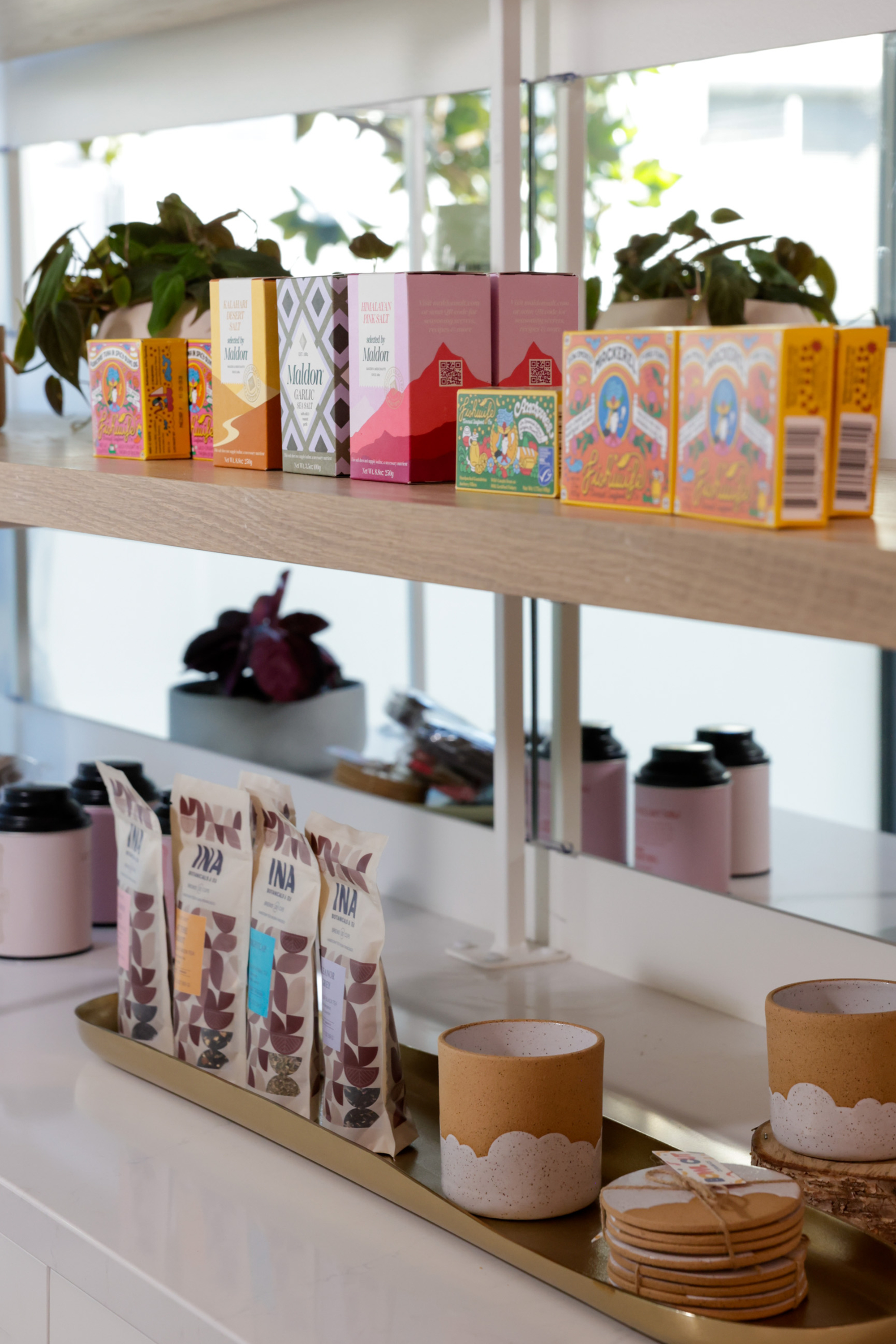 The image shows a wooden shelf with colorful packages and small plants. Below, a counter holds packaged snacks, ceramic cups, and a stack of coasters.