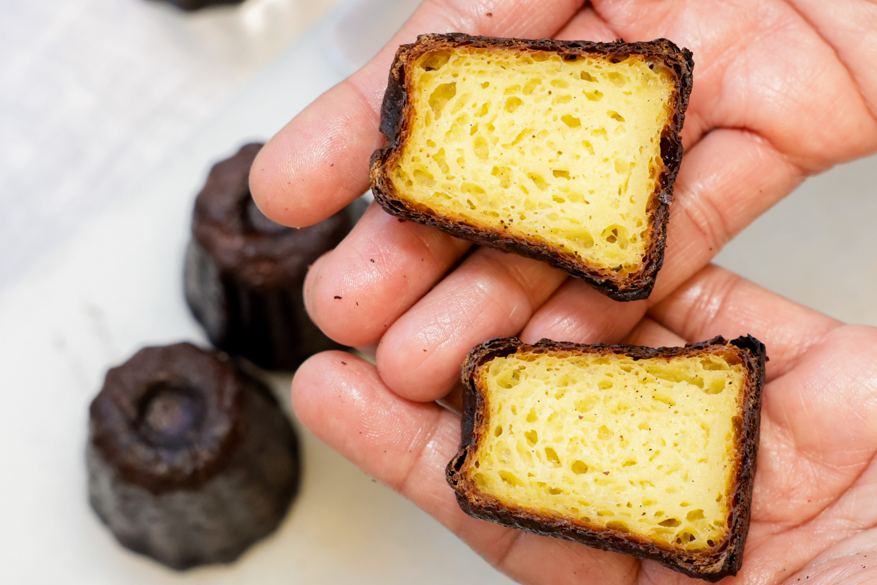 A person holds a halved pastry with a caramelized brown crust and a soft, airy yellow interior, with two uncut pastries in the background.