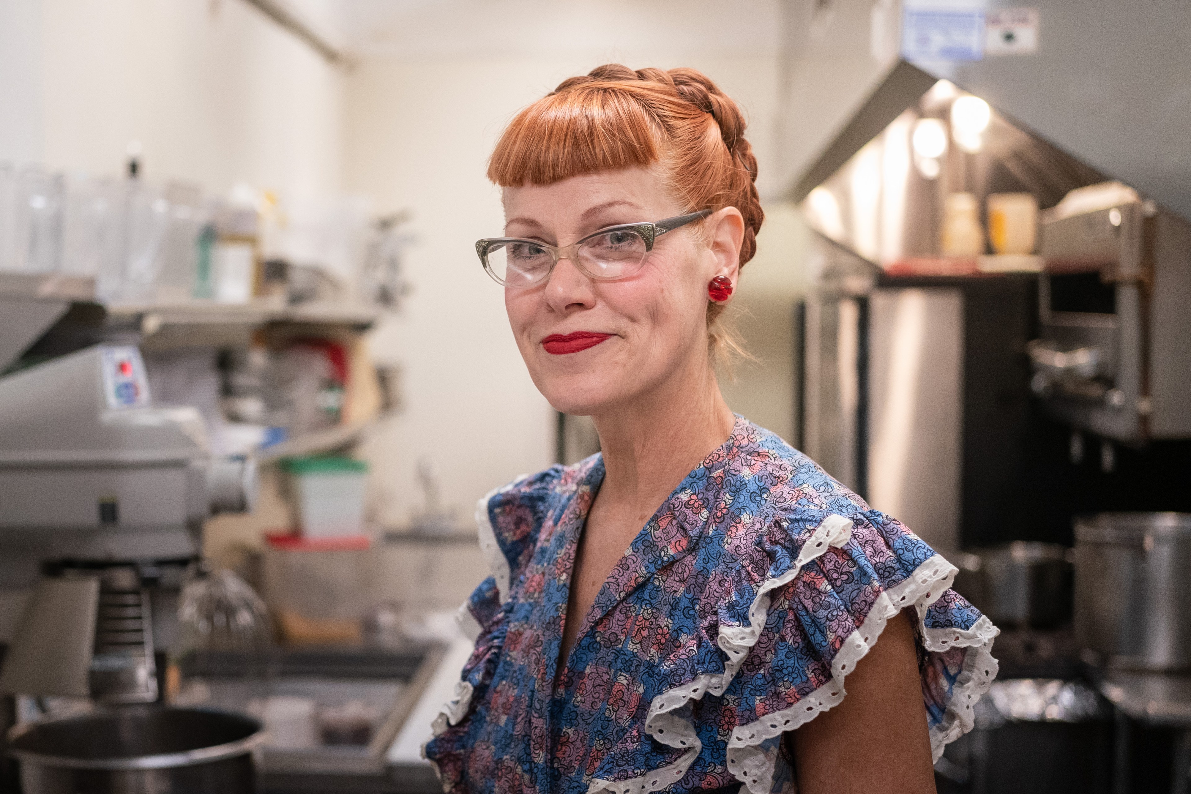 A woman with red hair in a braid, wearing glasses and a floral dress, smiles in a kitchen setting with various cooking equipment in the background.