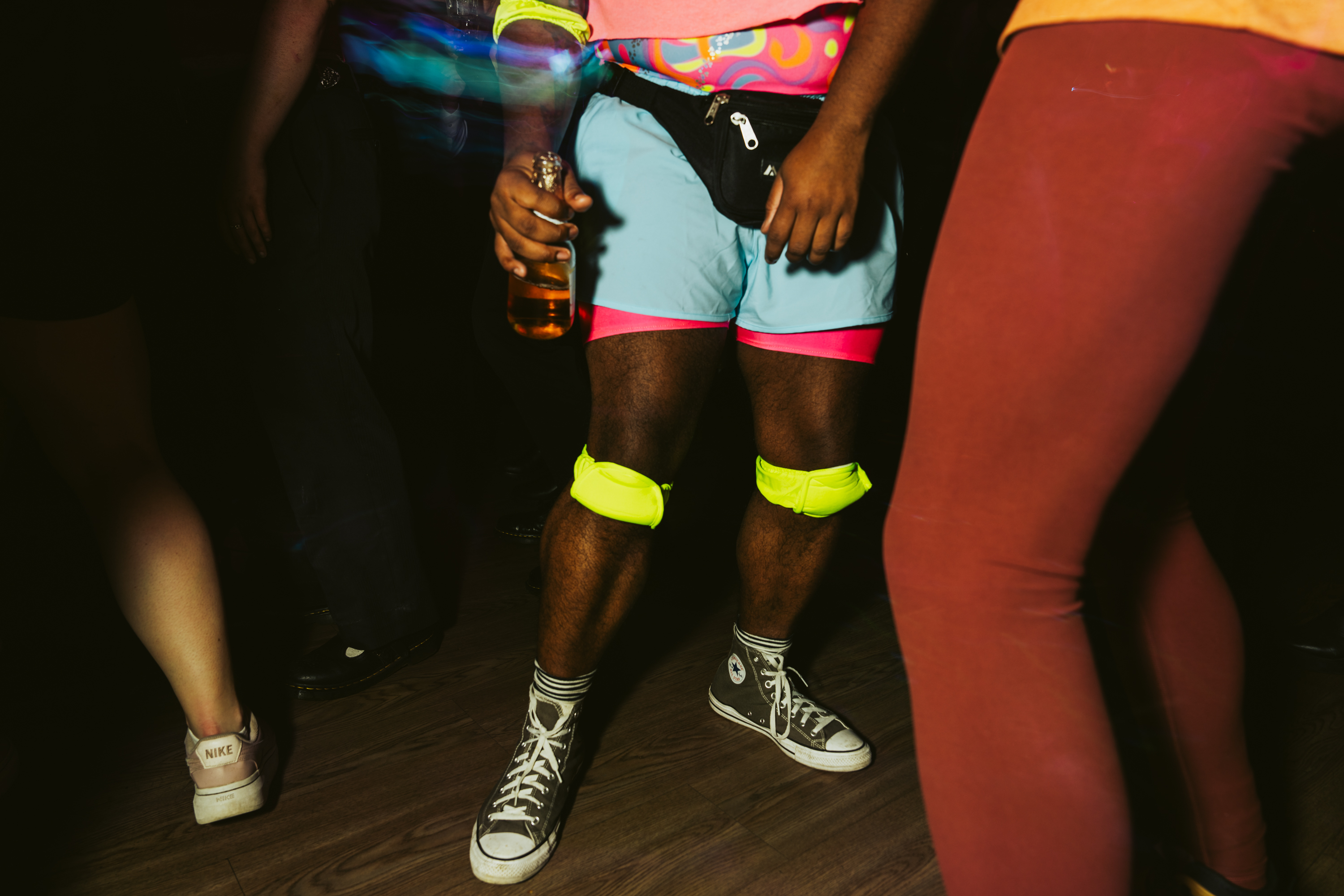 People are dancing in a dimly lit room. A person in bright shorts and neon kneepads holds a drink, surrounded by others in casual attire, including sneakers.