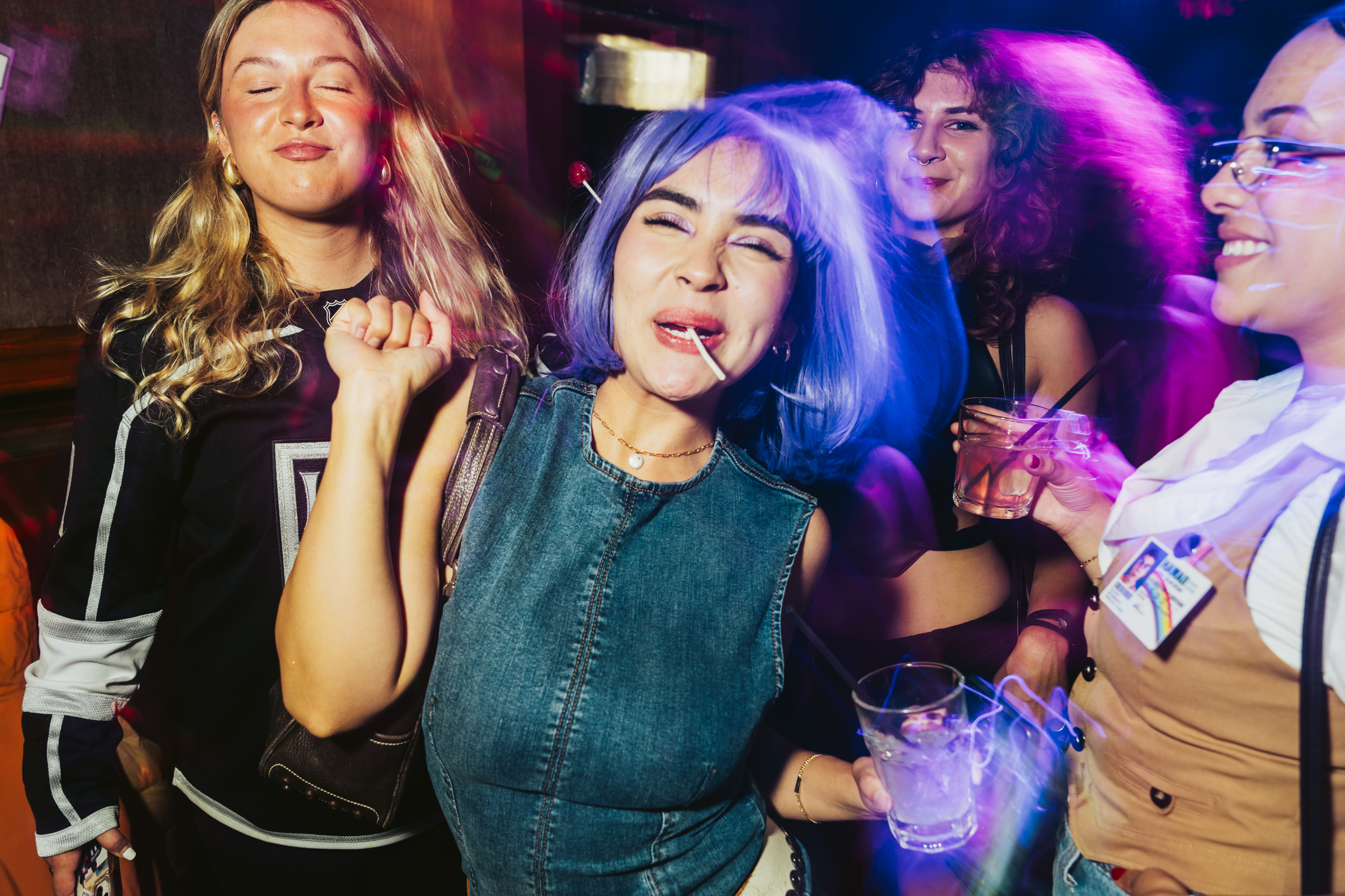 A group of people are having fun in a vibrant setting. The central figure, with blue hair and a lollipop, is dancing and holding a drink, surrounded by friends.