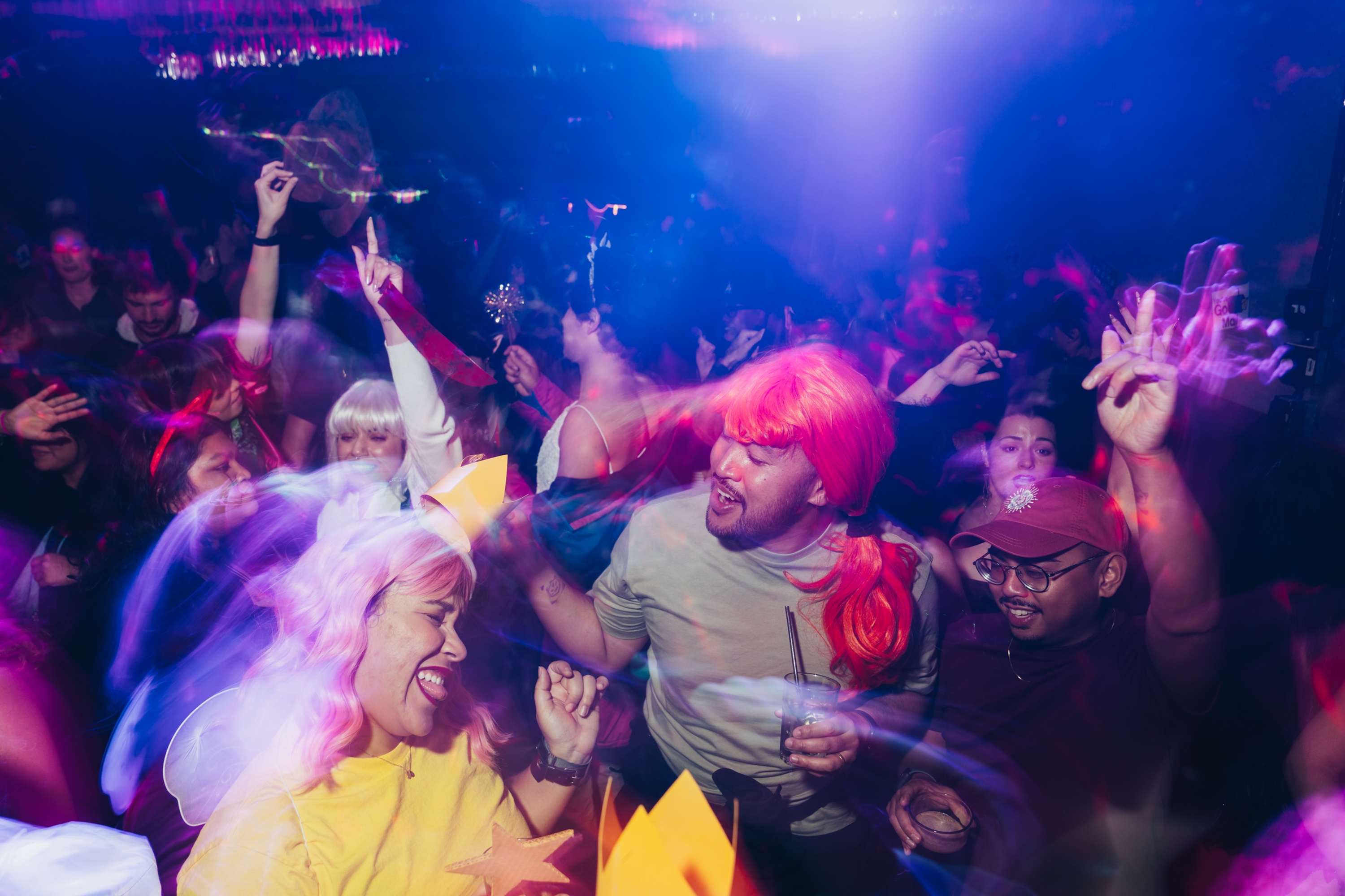 A joyful crowd in colorful costumes dances under vibrant lights, creating a lively atmosphere at a party or celebration.