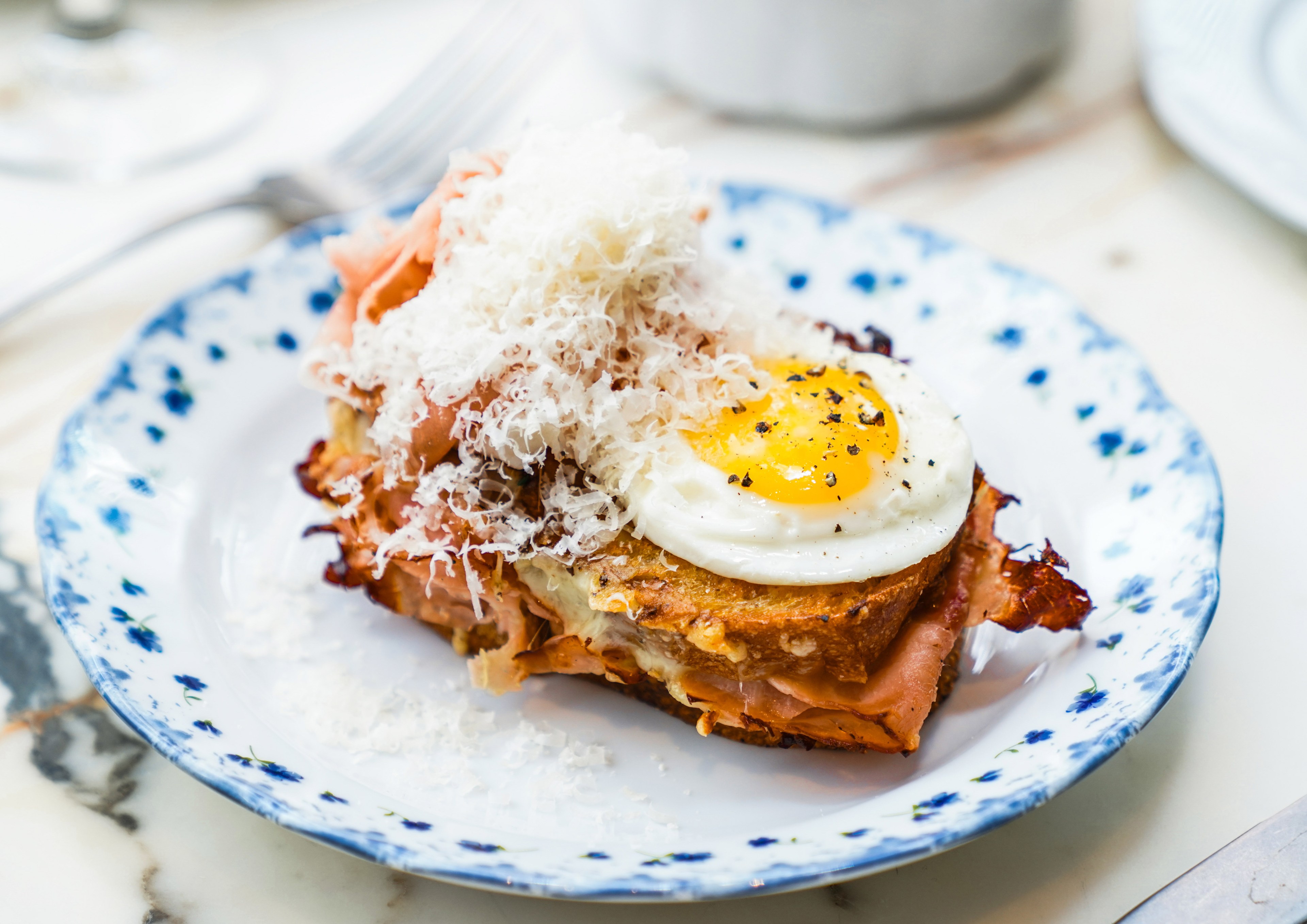 A plate with a toasted sandwich layered with thin slices of ham, a fried egg with pepper, and a generous topping of grated cheese, all on a decorative plate.