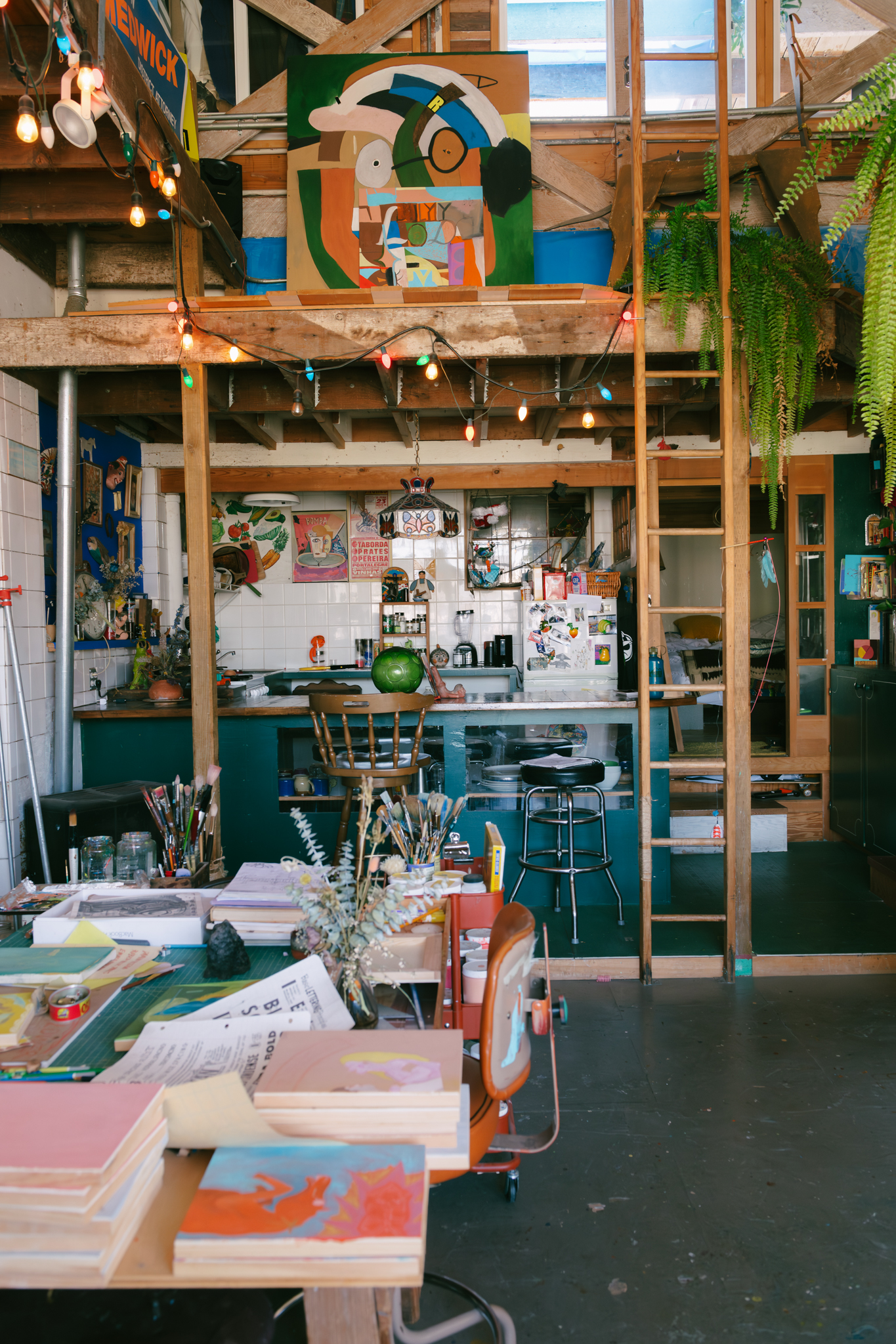 A vibrant art studio with colorful paintings, brushes, and art supplies on tables. String lights hang above, and there's a small kitchen area in the back.