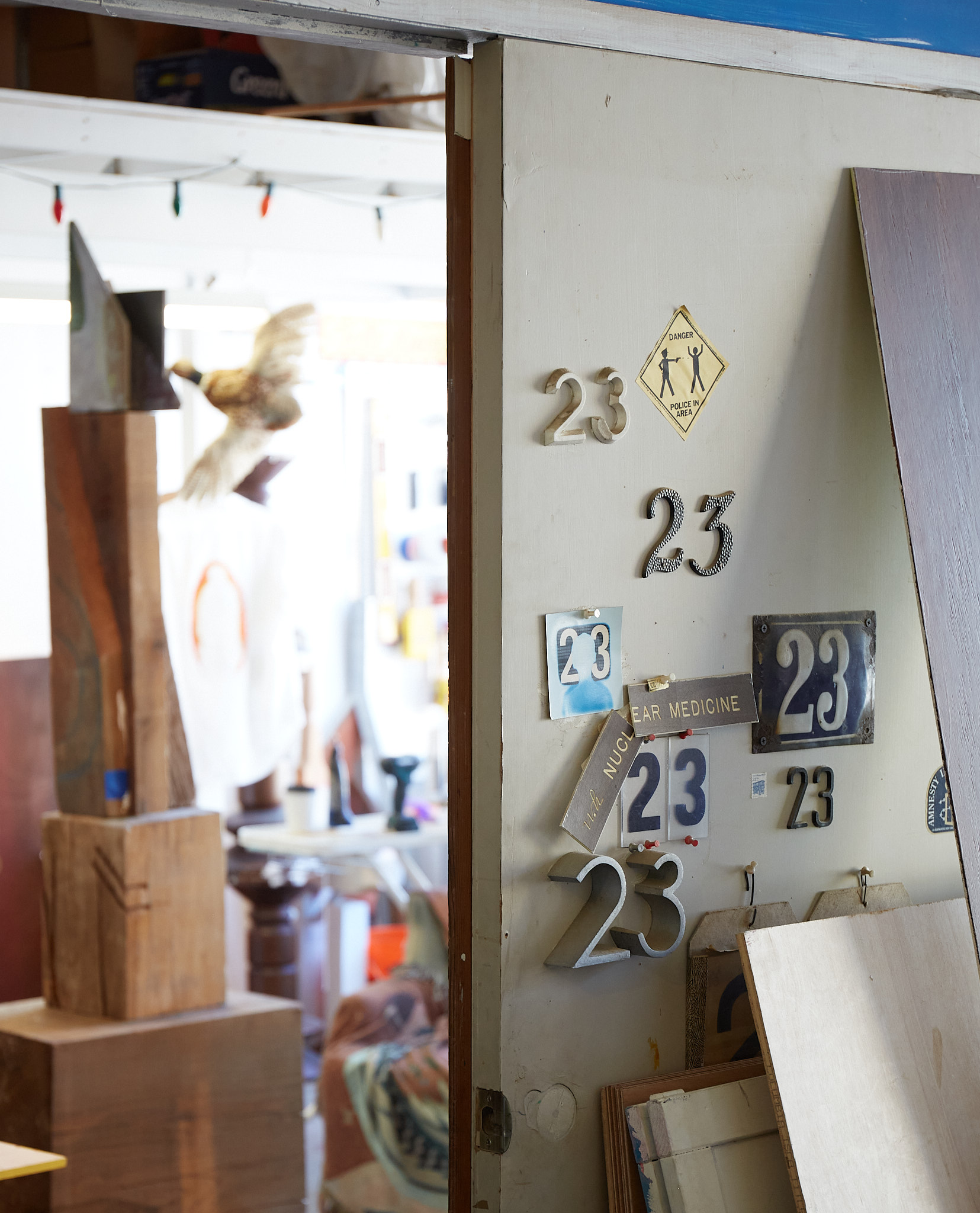 A door is adorned with various &quot;23&quot; signs and a &quot;Danger! Police in area&quot; notice. The room beyond is cluttered, with wooden blocks and a person partially visible.