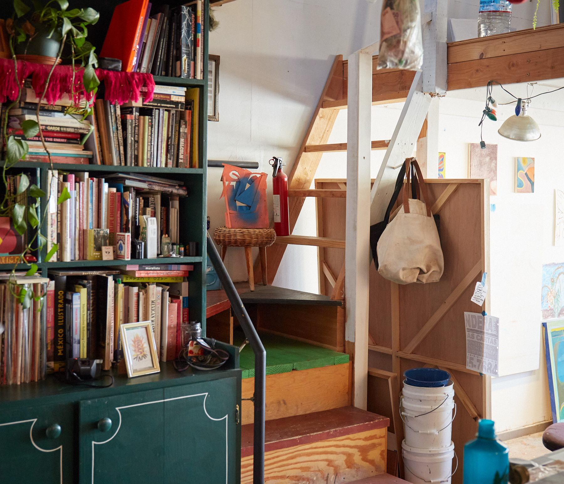 The image shows a cozy room with a bookshelf full of books and plants, a staircase with bags hanging, and buckets stacked underneath, creating an artistic vibe.