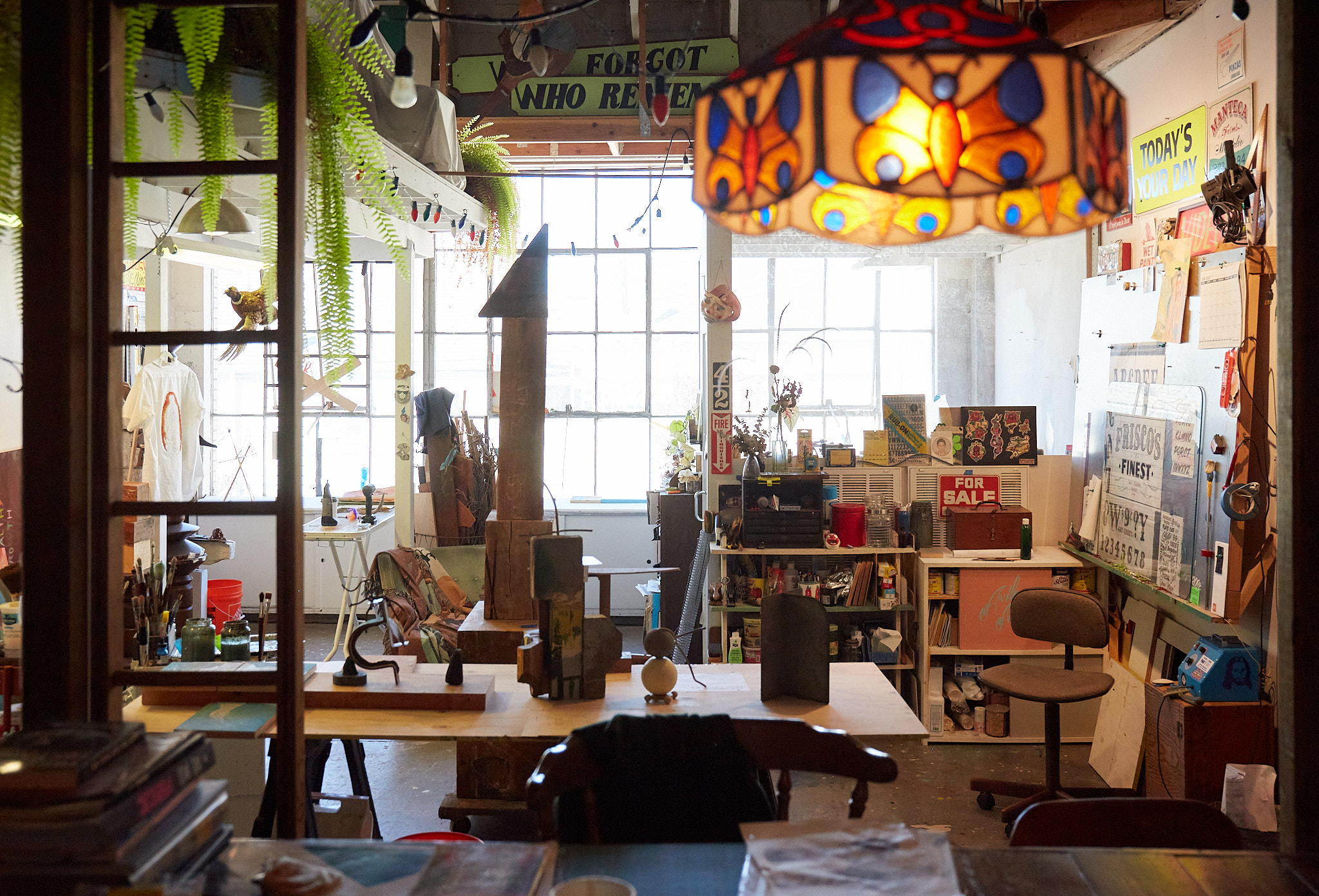 A cluttered, creative workshop with wooden furniture, plants, bright hanging lamp, art supplies, and posters. Natural light streams through large windows.