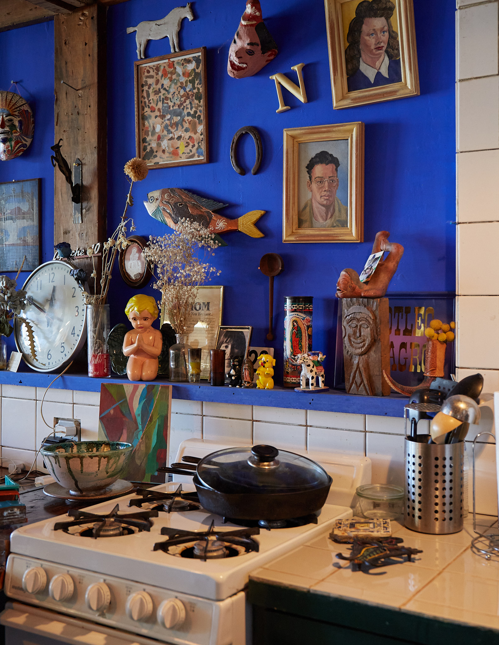 A kitchen with eclectic decor includes colorful masks, paintings, and a fish sculpture on blue walls. A stove with kitchen utensils and a pot sits below.