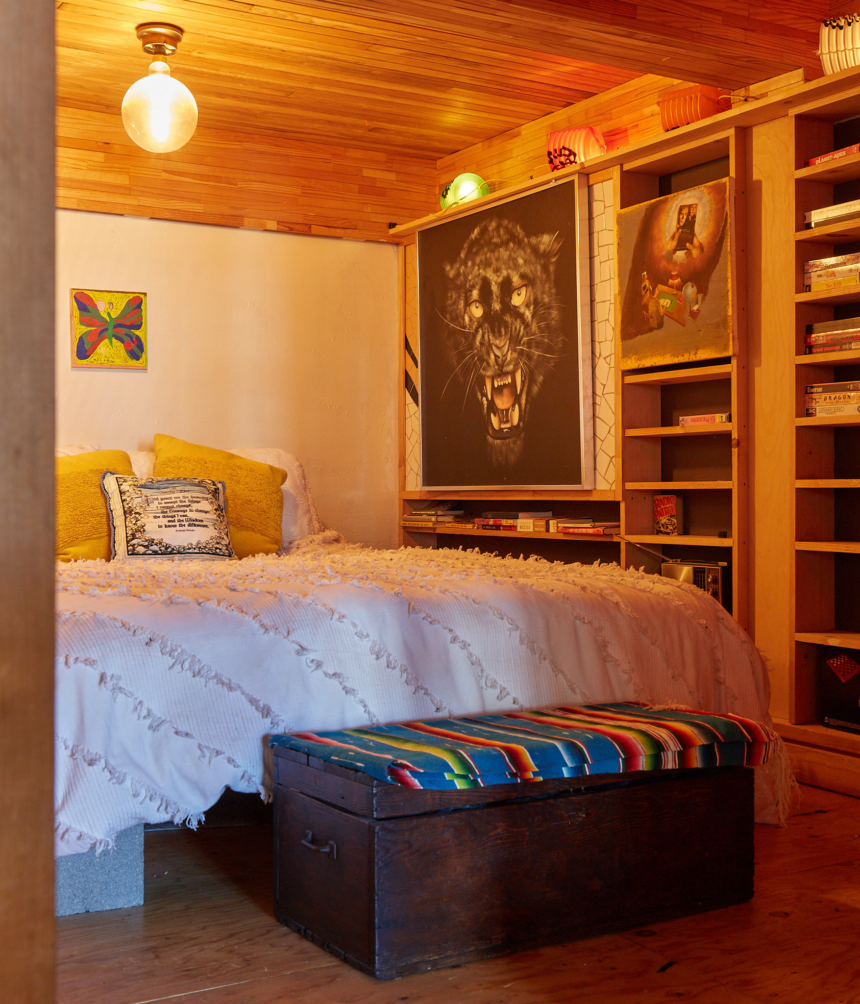 A cozy room with a bed featuring yellow pillows and a fluffy throw. A bold panther painting hangs above a bookshelf filled with various books. A colorful bench sits at the foot.