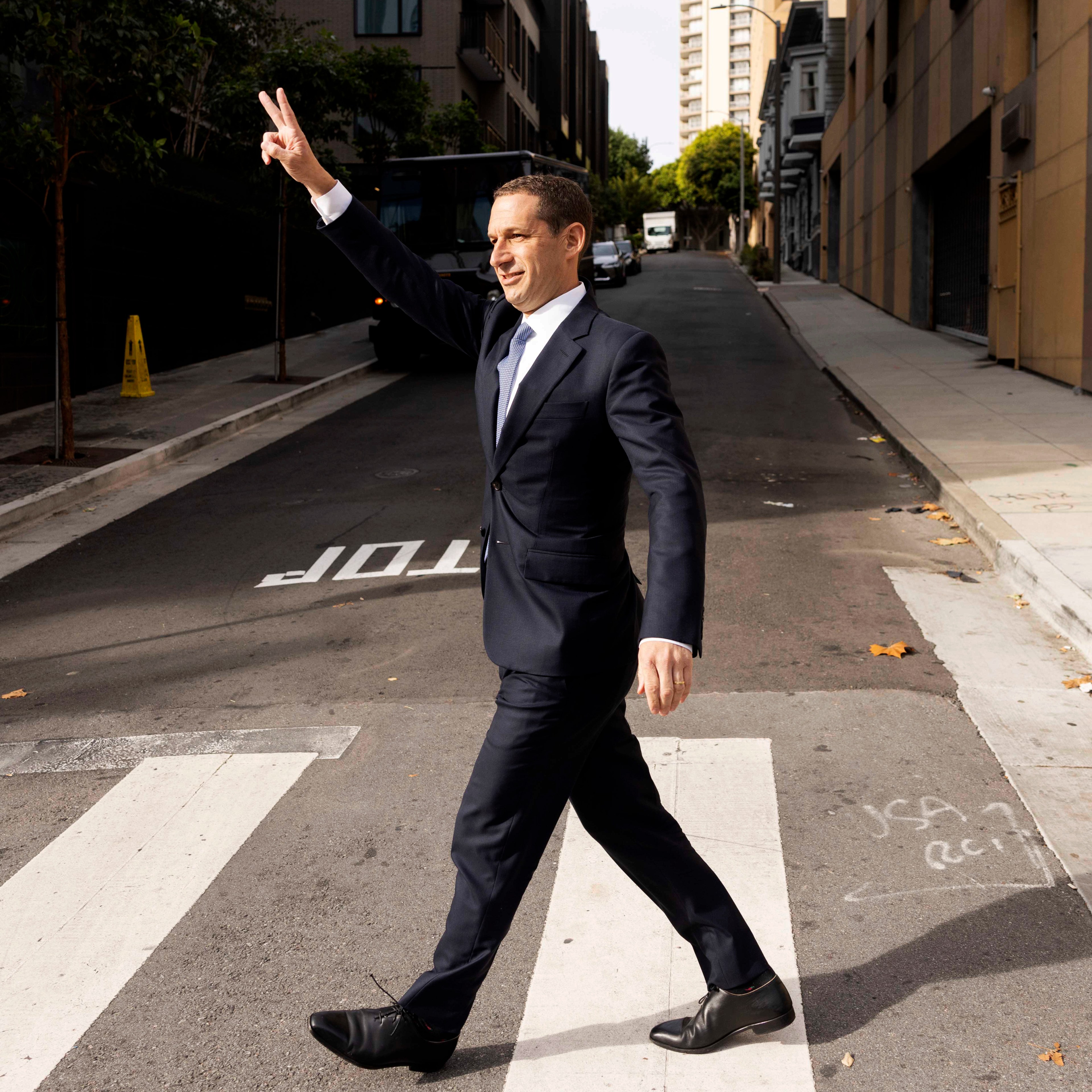 A man in a suit walks confidently across a crosswalk on a city street. He's raising his hand in a peace sign while smiling. The street is empty and lined with buildings.