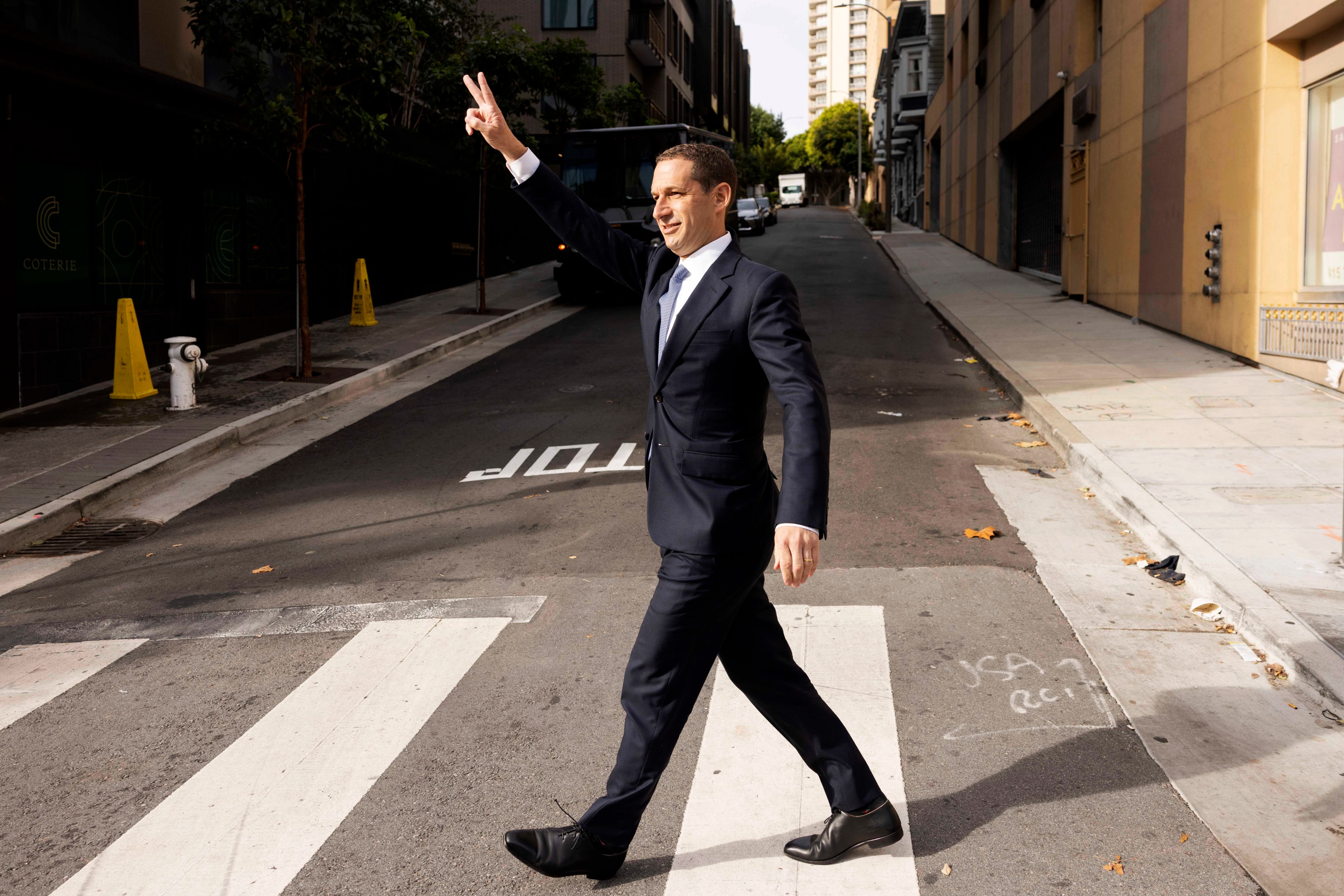 A man in a suit walks confidently across a crosswalk on a city street. He's raising his hand in a peace sign while smiling. The street is empty and lined with buildings.