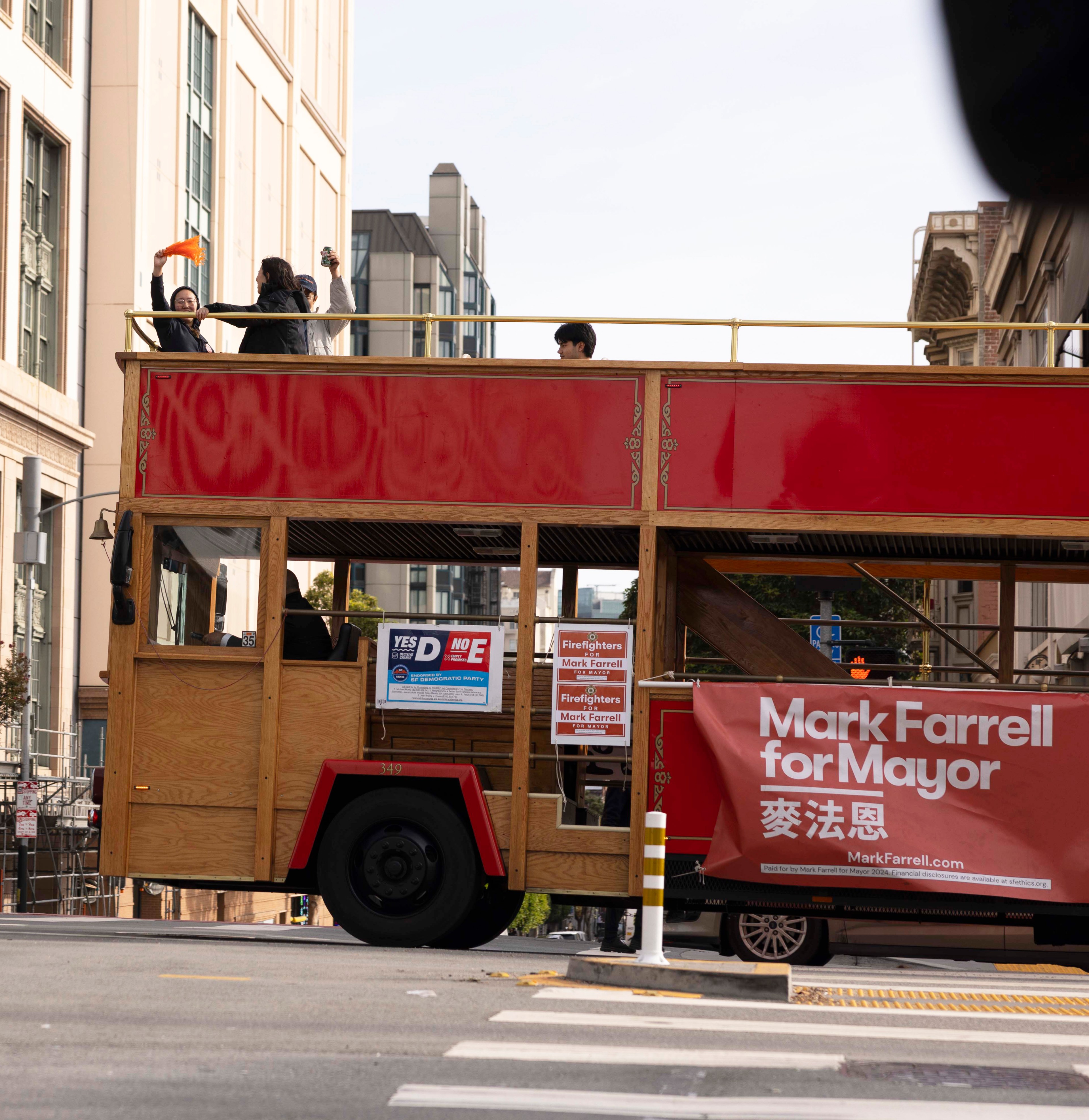 A double-decker bus with campaign signs for &quot;Mark Farrell for Mayor.&quot; People on the upper deck wave while the bus navigates an urban street.