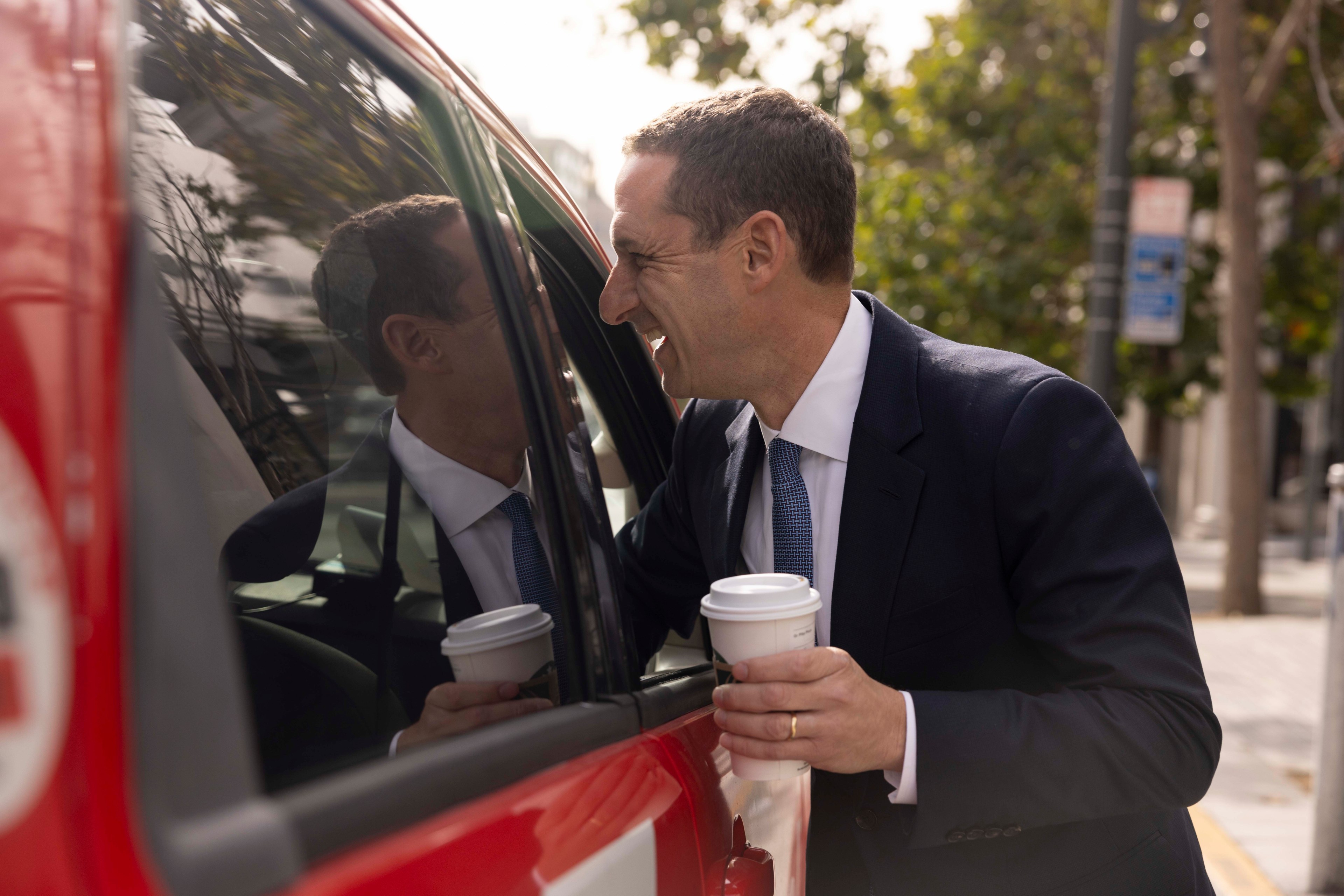 A man in a suit holds a coffee cup while leaning toward a red vehicle, smiling at his reflection in the window on a sunny day with trees in the background.